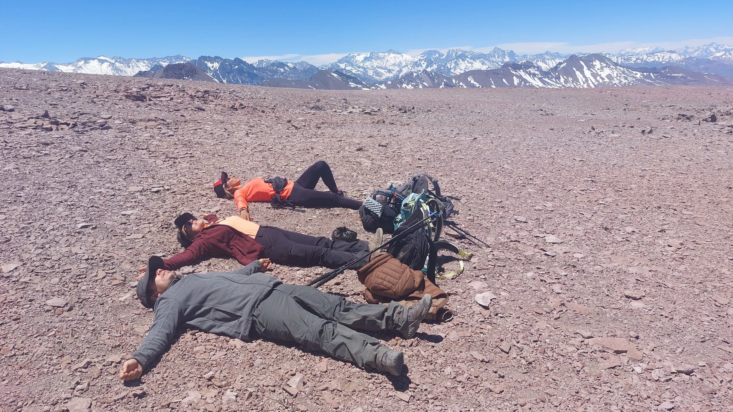 Tres personas descansando en una montaña rocosa con nieve de fondo, rodeadas de equipo de senderismo y con un paisaje de montañas nevadas y un cielo despejado.