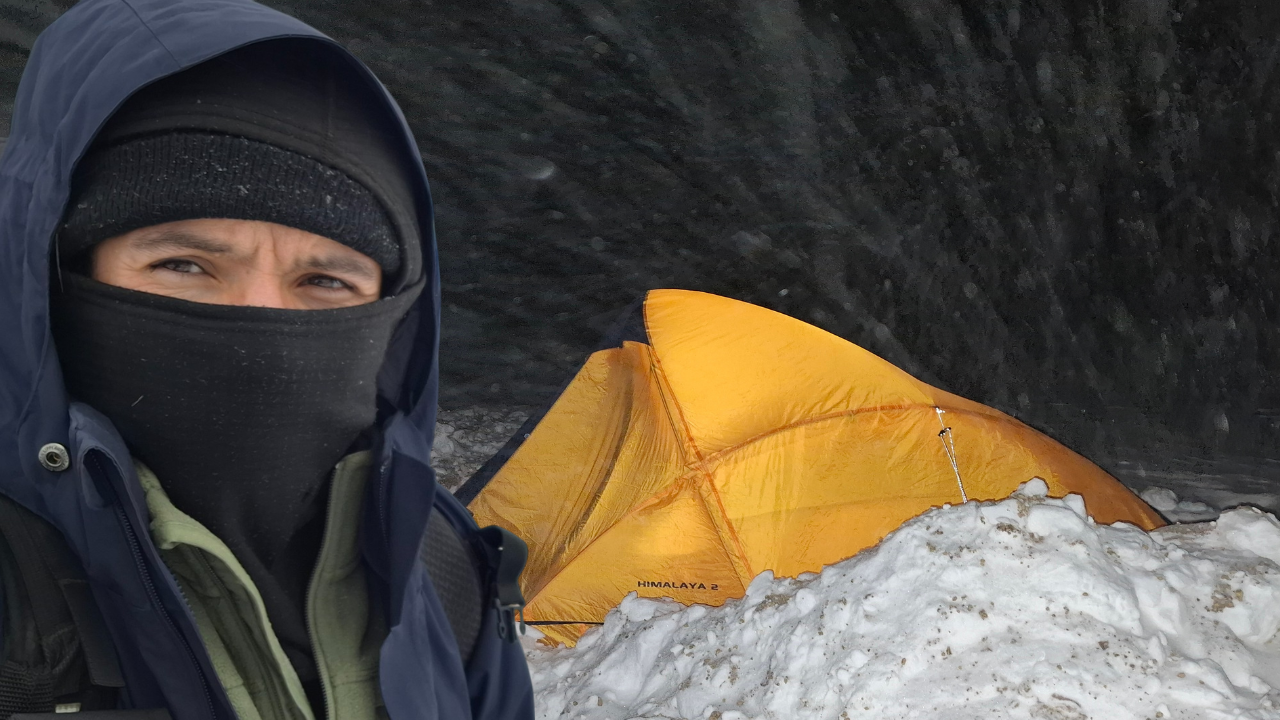 Persona con ropa de abrigo en un ambiente de nieve, al fondo una tienda de campaña color naranja y cumbres nevadas.