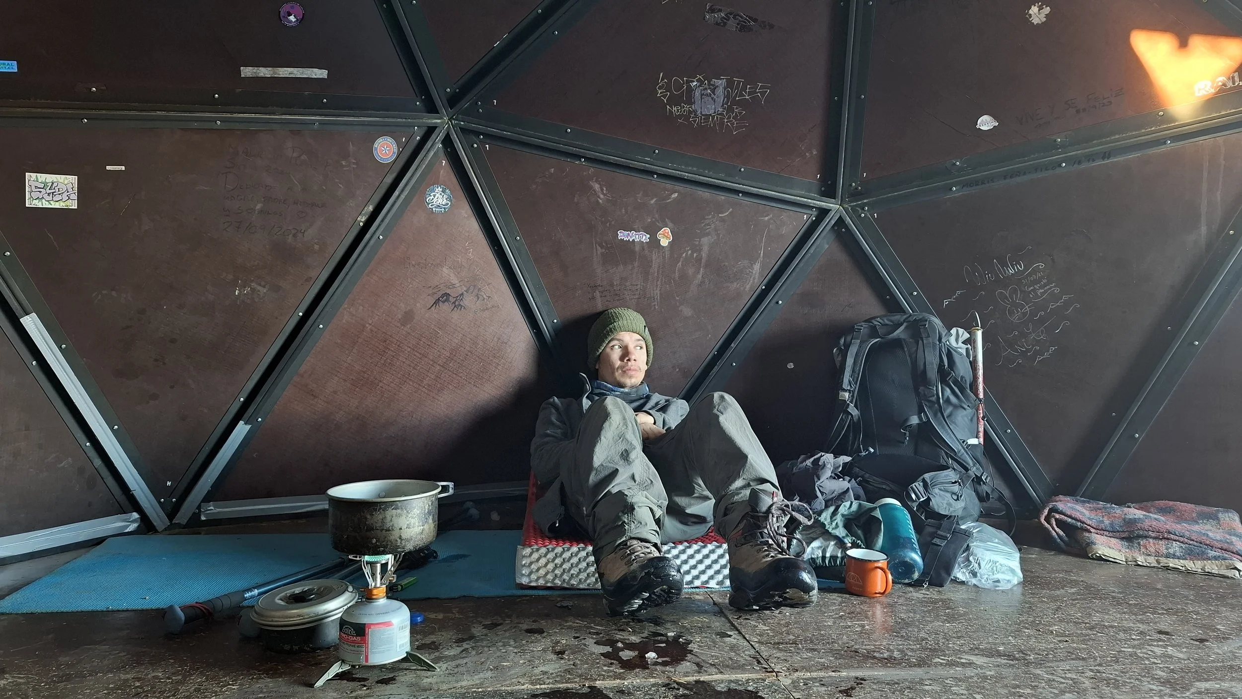 Persona descansando en una cabaña con estructura de marco metálico y paredes de madera, rodeada de objetos como mochila, carpa, utensilios de camping y graffiti en el techo.