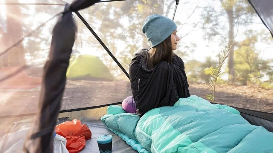 Una mujer sentada en una tienda de campaña en un entorno natural, con vista a árboles y luz de día, vestida con gorro azul y chaqueta negra, rodeada de una almohada, una botella y equipo de camping.