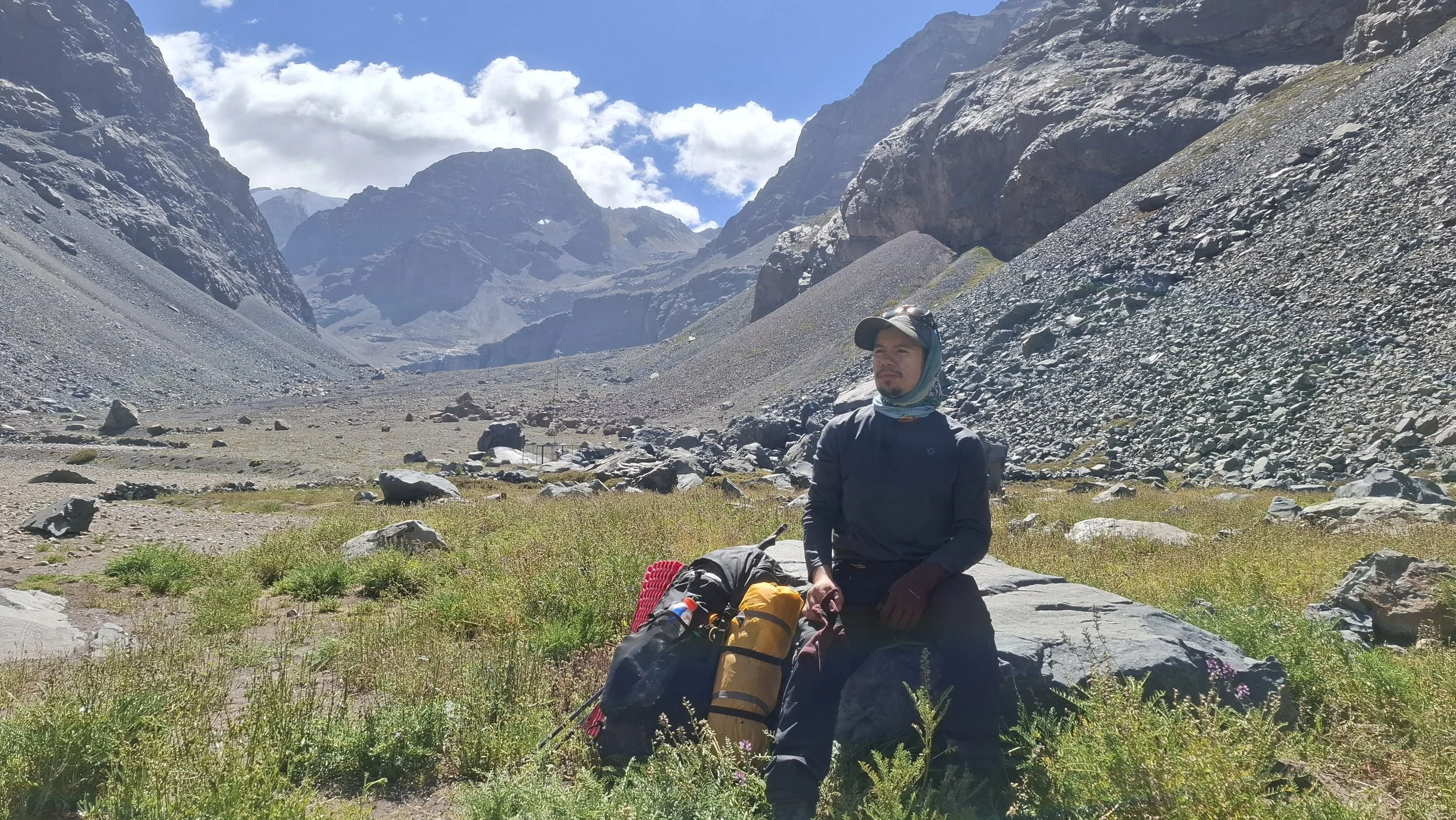 Persona descansando en un paisaje montañoso con rocas, vegetación y cielo con nubes.