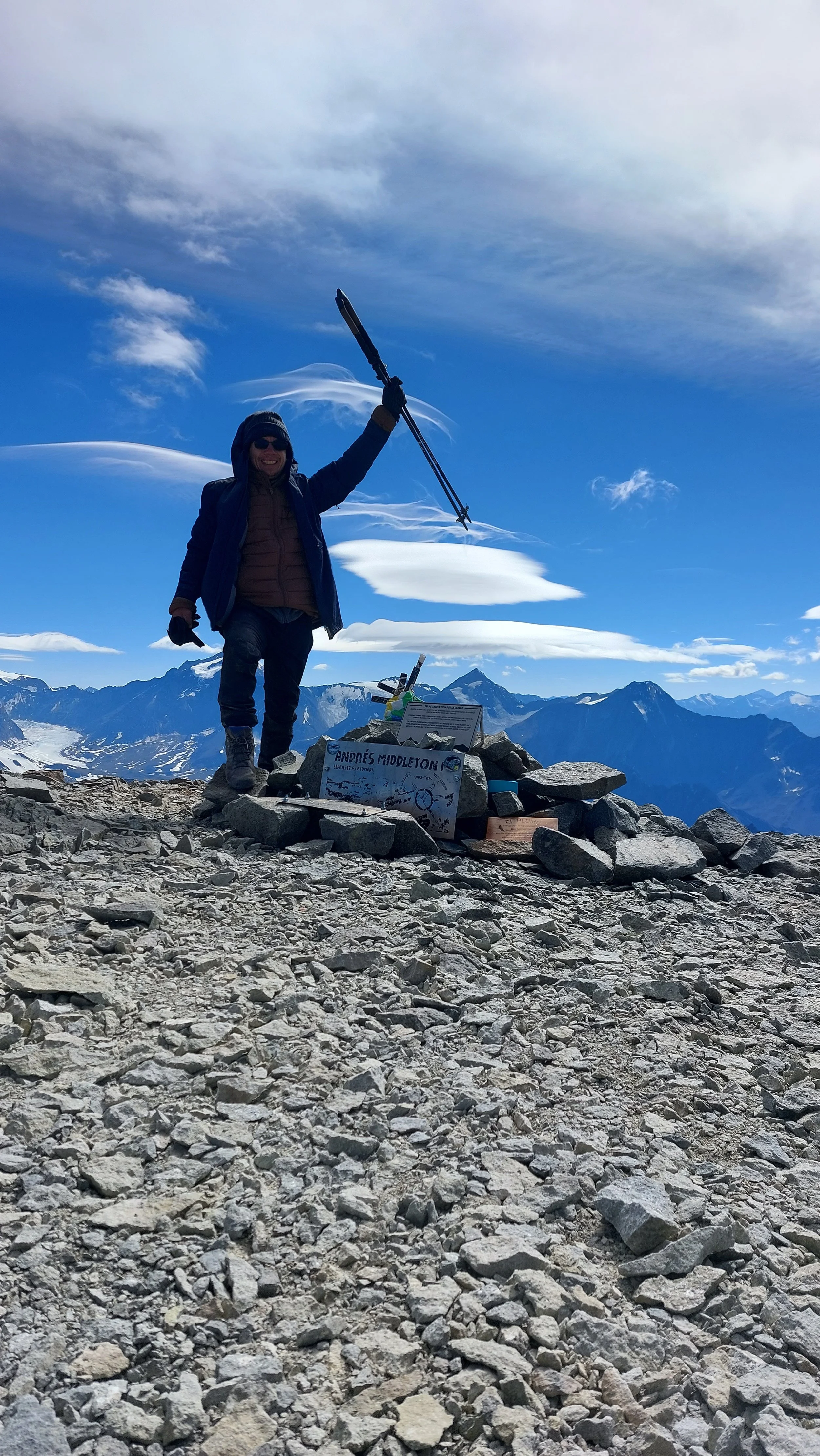Persona en la cima de una montaña, celebrando con una pica en la mano y sonriendo. Está rodeada de rocas y tiene un cartel que dice "Andrés Middleton". Delante, hay una pila de piedras y un panel informativo, con montañas y un cielo despejado en el fondo.