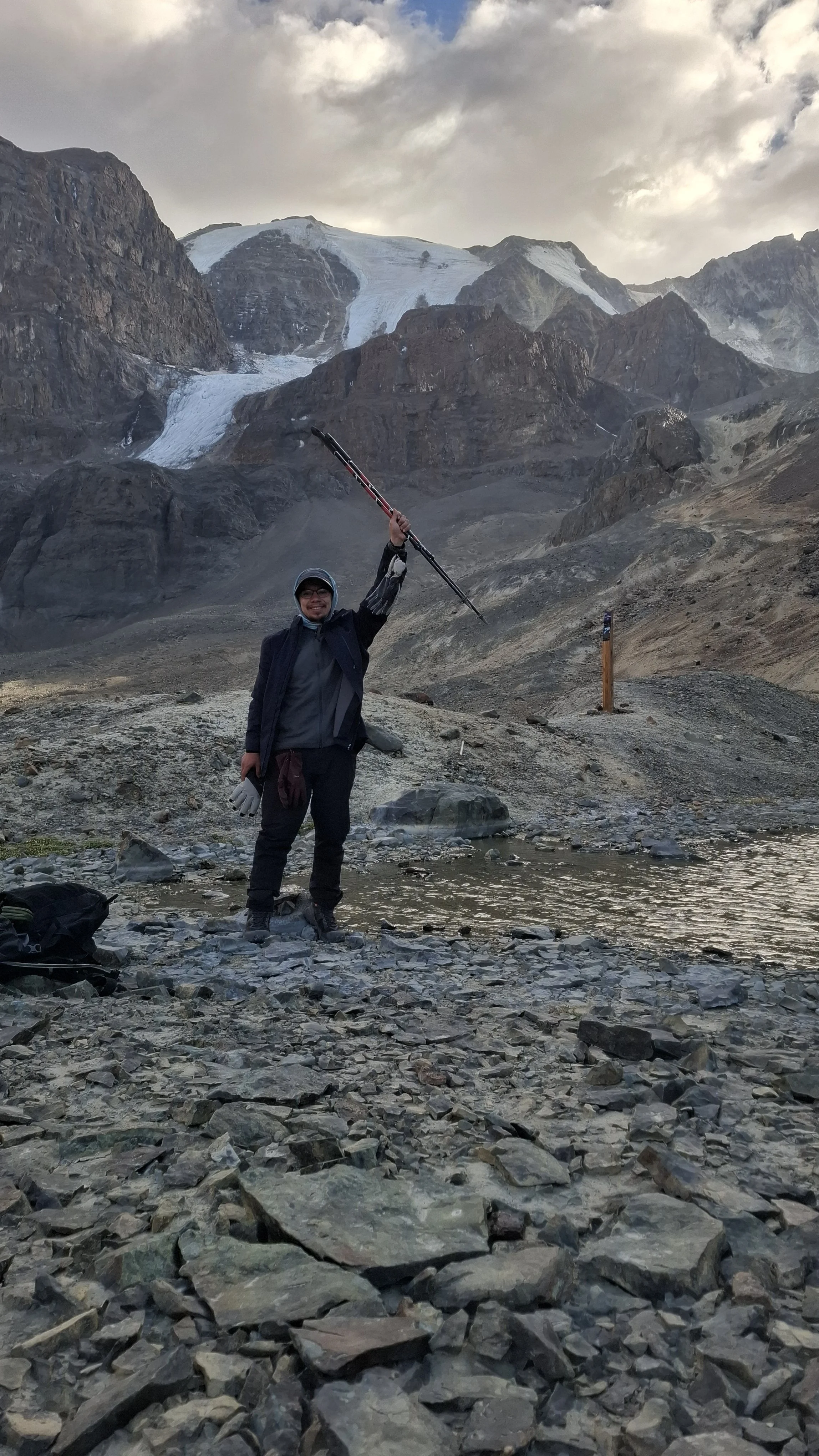 Persona con casco y ropa de abrigo levantando un bastón en un paisaje montañoso con glaciares, rocas y un río pequeño.