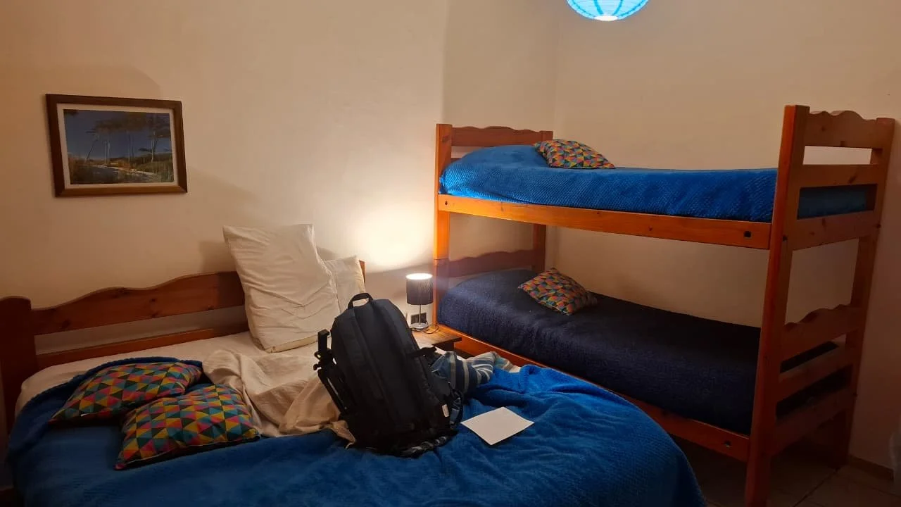 Habitación con cama doble y literas de madera con colchones azules, almohadas con fundas coloridas, mochila negra en la cama, lámpara y cuadro en la pared