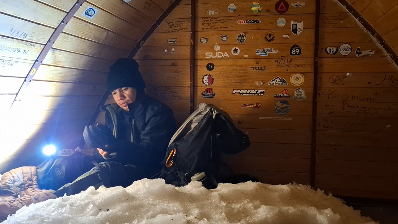 Persona en una cabaña de madera usando ropa de abrigo, sosteniendo una linterna y revisando su teléfono, con un saco y otros objetos a su lado, paredes adornadas con calcomanías y notas, y nieve en el suelo.