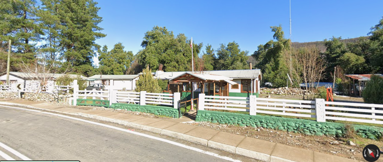 Casa con paredes blancas y detalles en verde, rodeada de árboles y cercada por una reja blanca, en un entorno rural con camino de asfalto en primer plano.