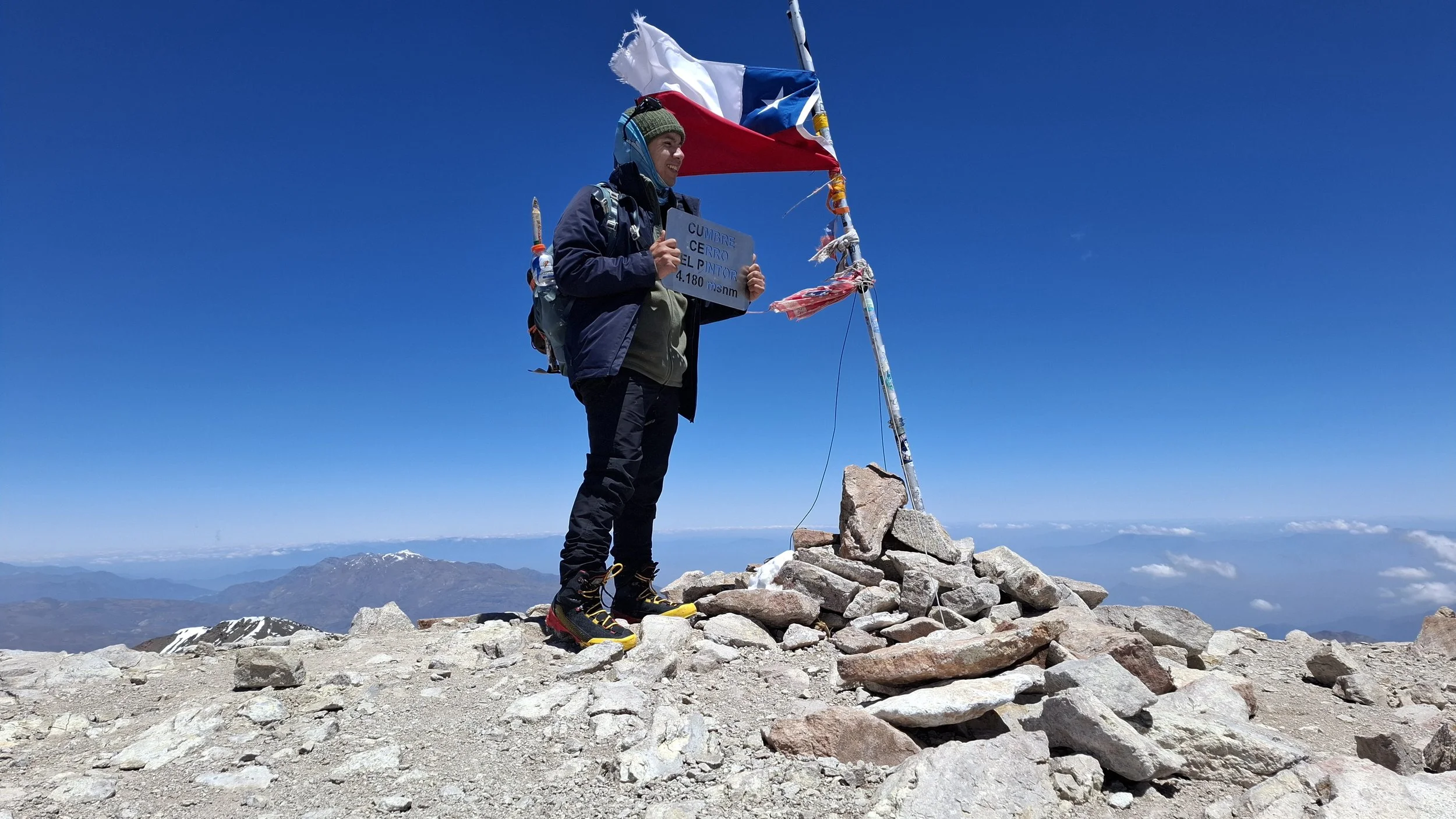 Persona en la cumbre de una montaña sosteniendo un cartel y una bandera chilena, rodeada de montañas y cielo azul.