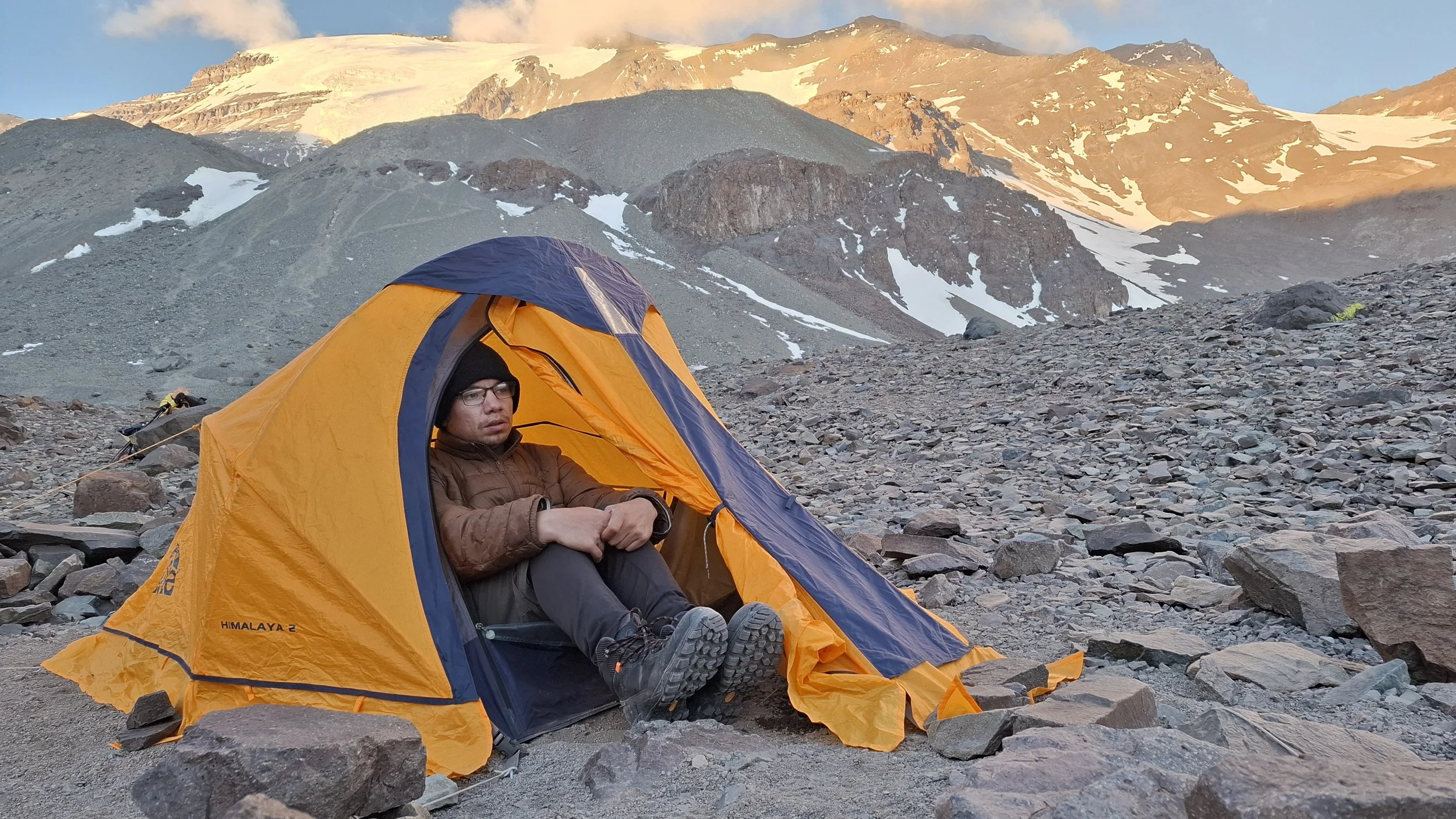Una persona sentada en la entrada de una tienda de campaña naranja en un paisaje montañoso con nieve y rocas, durante el amanecer o atardecer.