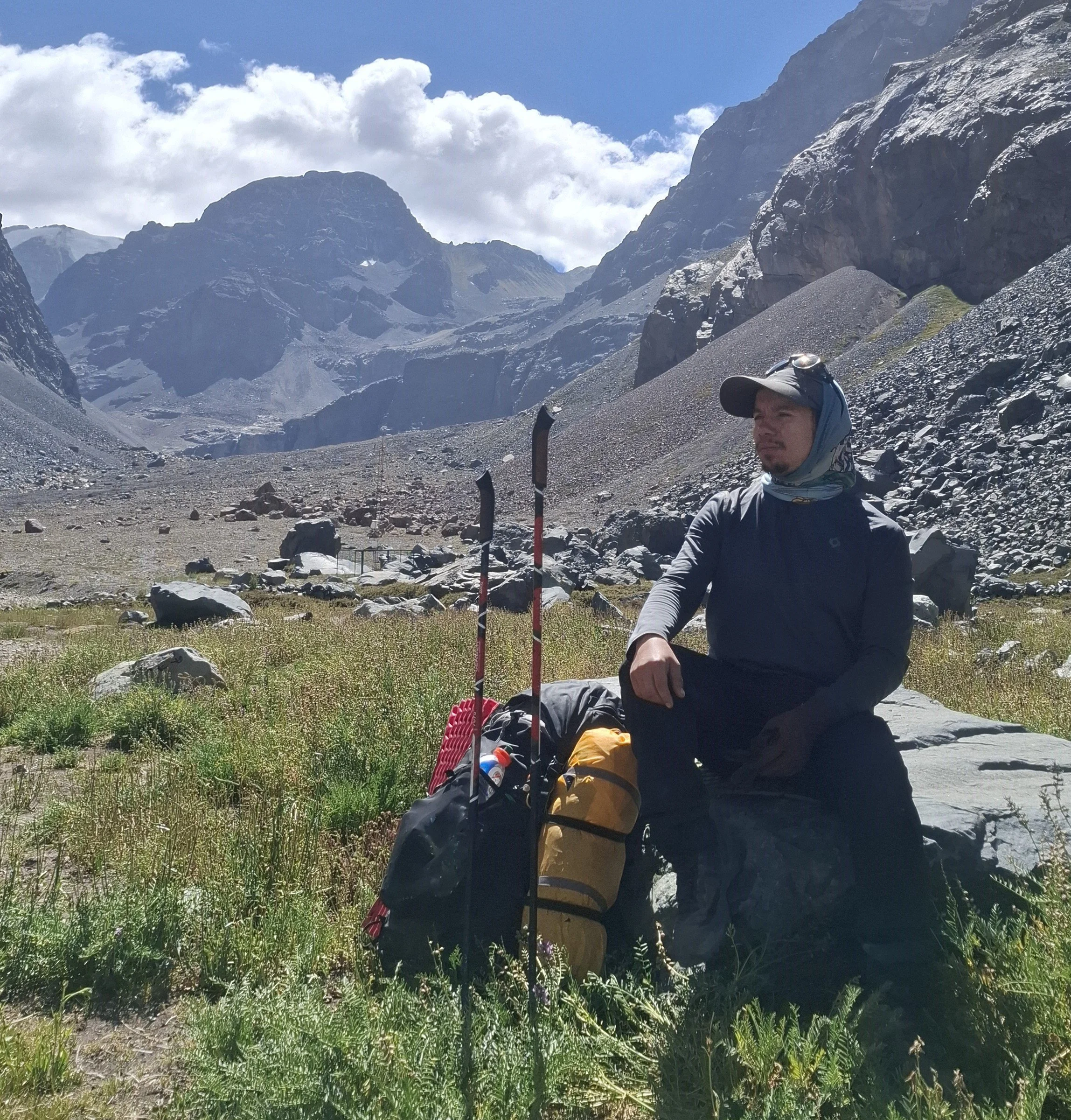 Persona con gorra y bufanda en montañas rocosas, paisaje de picos y nubes, equipo de senderismo con mochila y bastón.