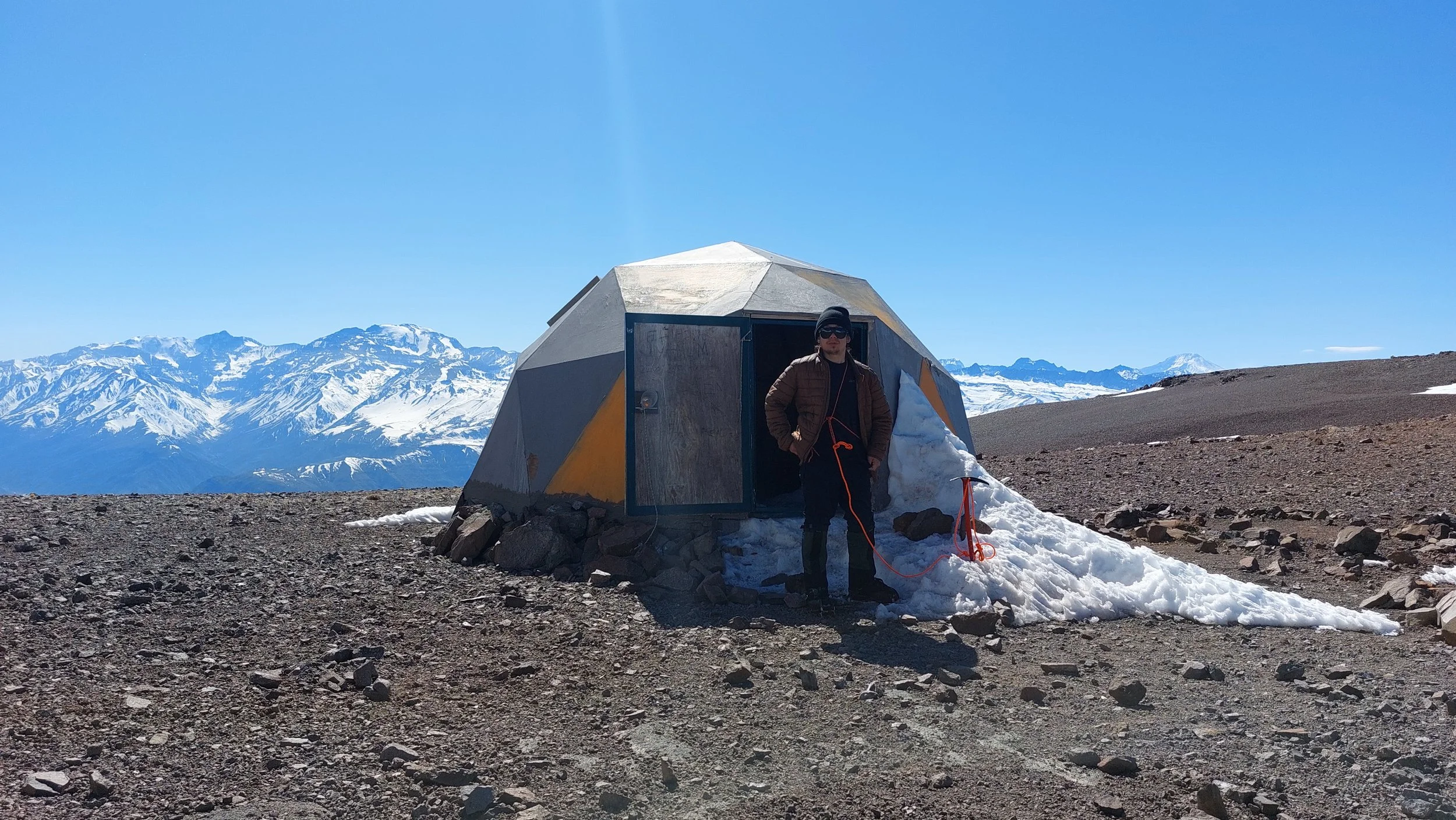Persona de pie afuera de una cabaña en una montaña nevada, con paredes metálicas y un paisaje de montañas con nieve al fondo.