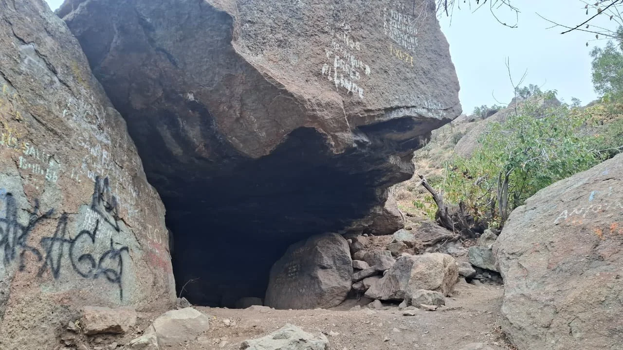 Entrada a una cueva en un área rocosa con grafiti en las rocas cercanas y vegetación en el fondo.