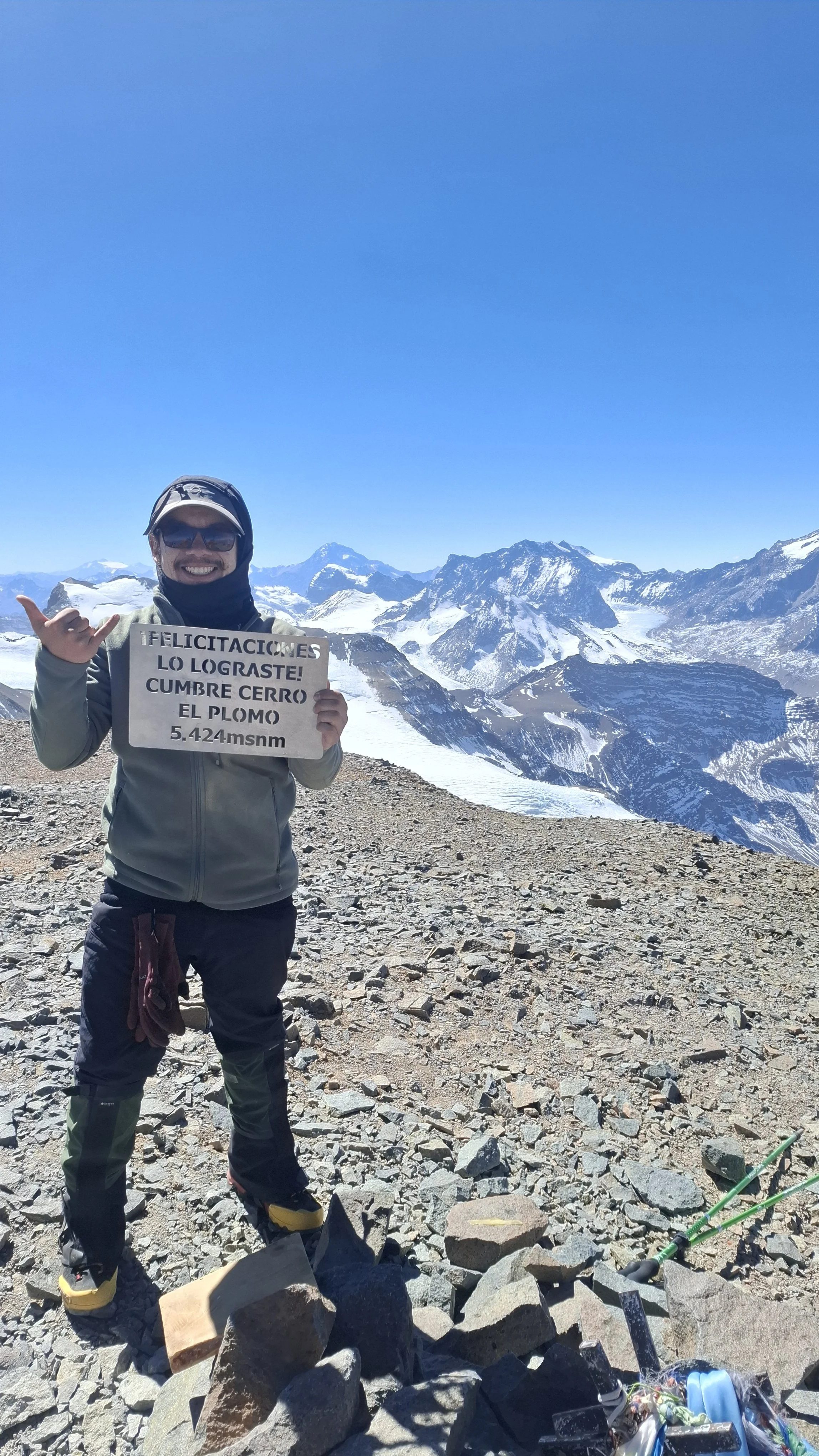 Persona en la cima de una montaña nevada sosteniendo un cartel que dice: '¡Felicitaciones lo lograste! Cumbre Cerro El Plomo 5.424 msnm con fondo de montañas cubiertas de nieve y un cielo azul claro.