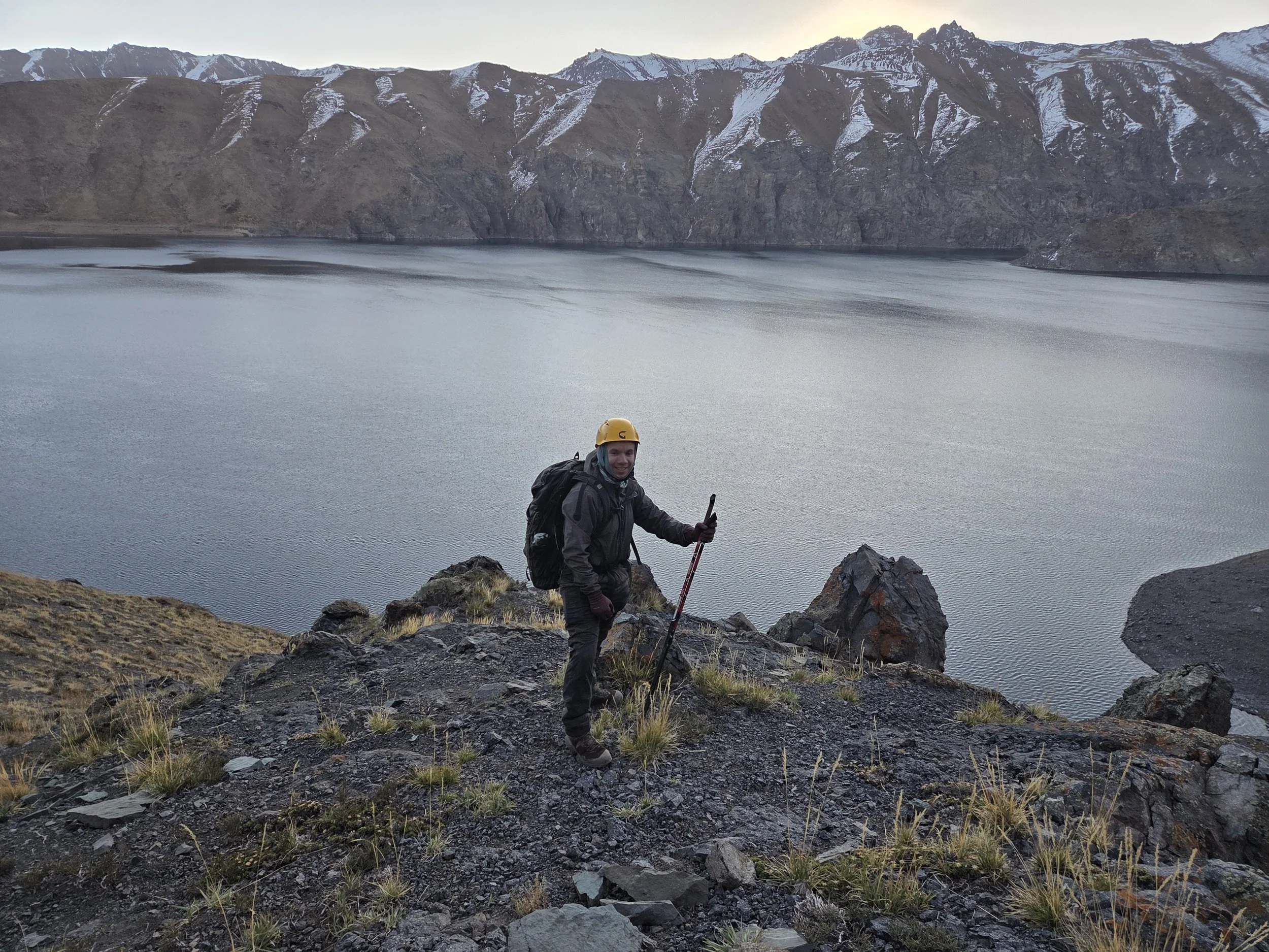 Persona con casco y mochila de pie en un paisaje montañoso junto a un cuerpo de agua, con montañas con nieve en el fondo.