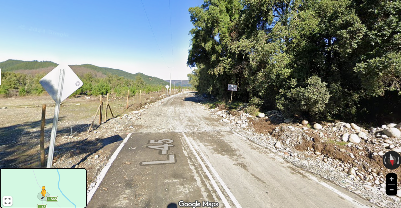 Camino rural en un ambiente montañoso con árboles grandes a la derecha y un letrero, con un camino de tierra y un cartel de limite de velocidad L-45.