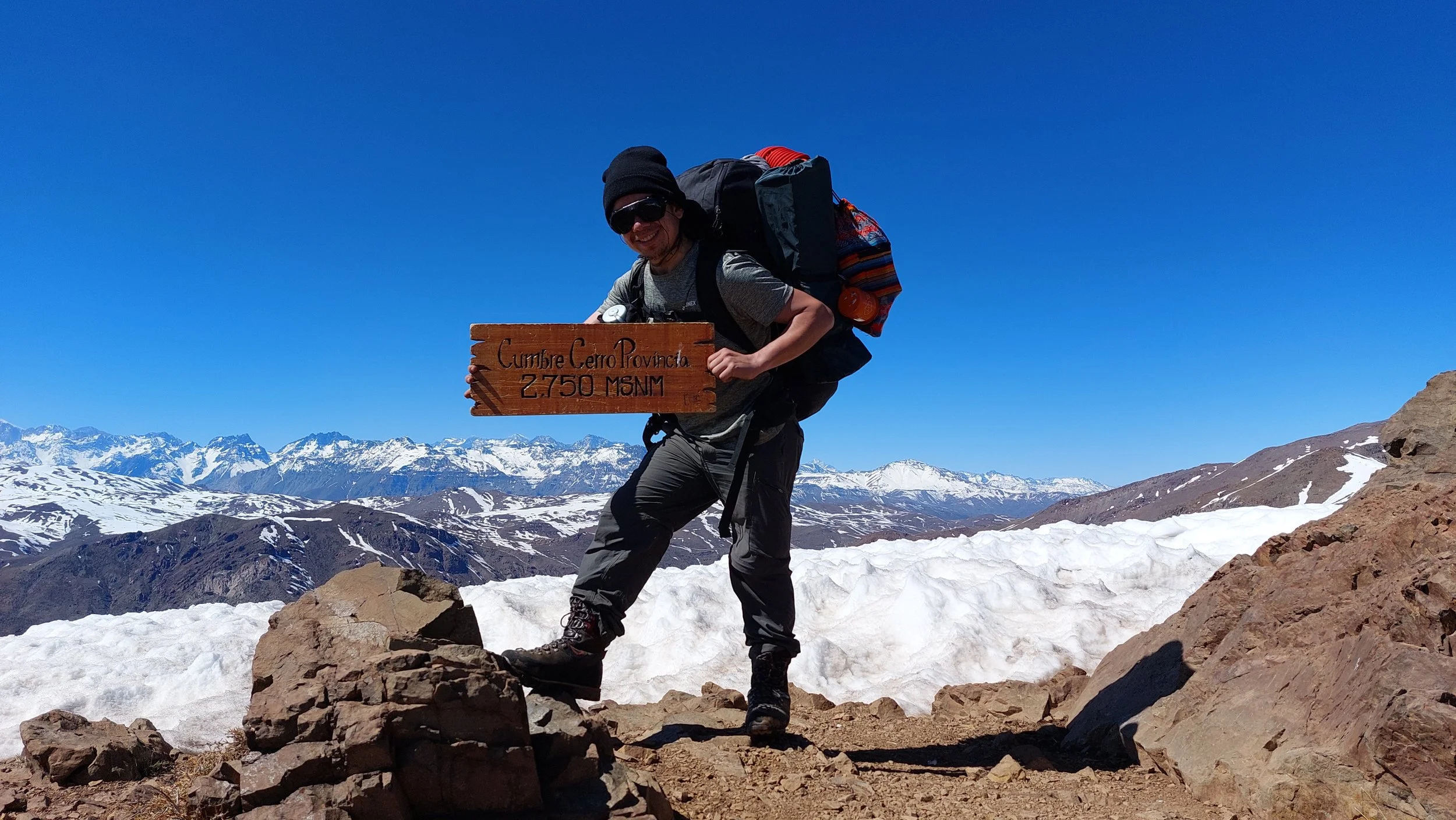 Persona en la cima de una montaña con nieve, sosteniendo un cartel que dice 'Cumbre Cerro Provincia 2750 MSNM', con un paisaje de montañas nevadas y un cielo despejado.