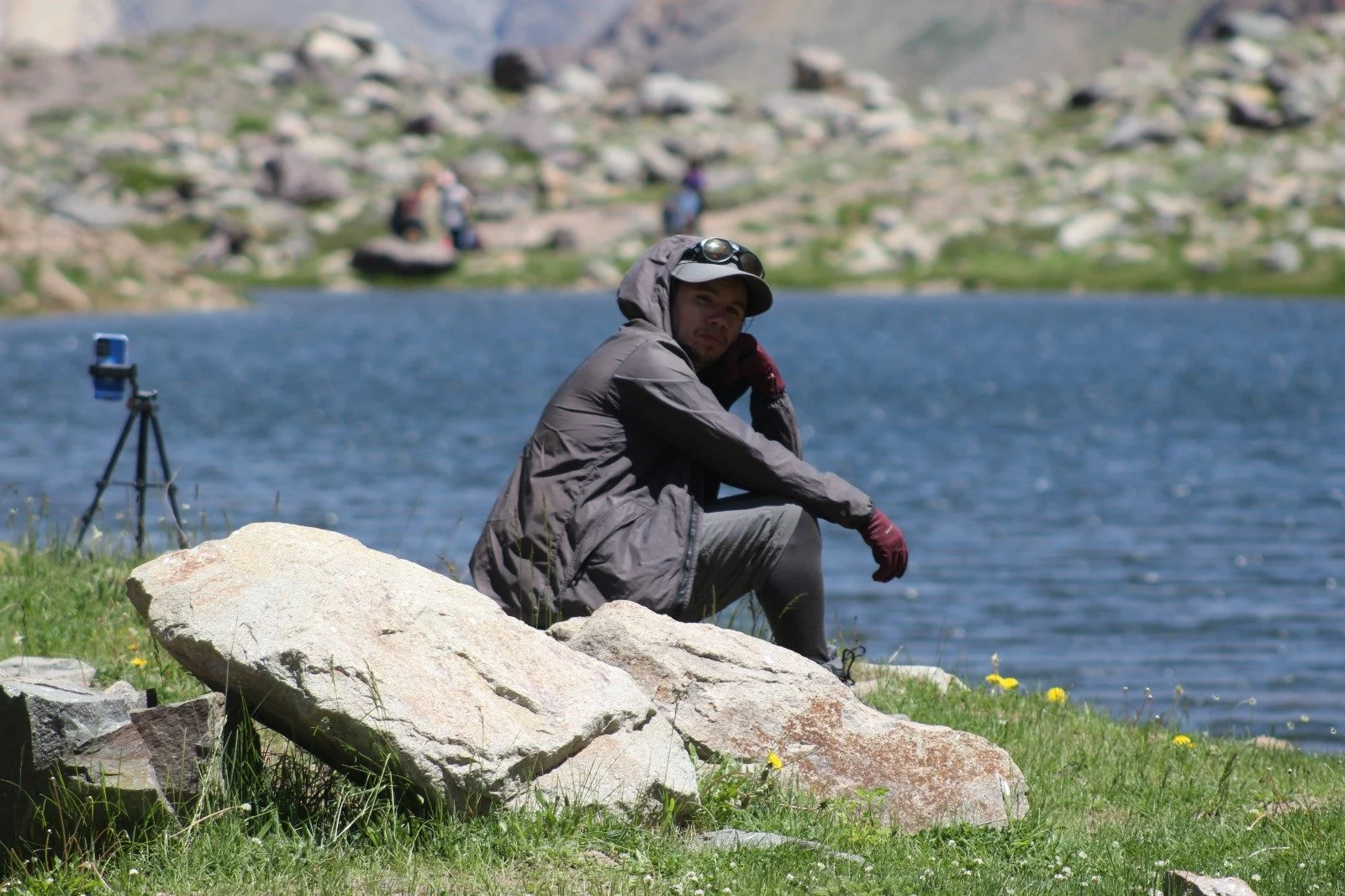 Persona sentada sobre rocas junto a un cuerpo de agua, con un fondo de montañas y otros visitantes, usando ropa para actividades al aire libre.