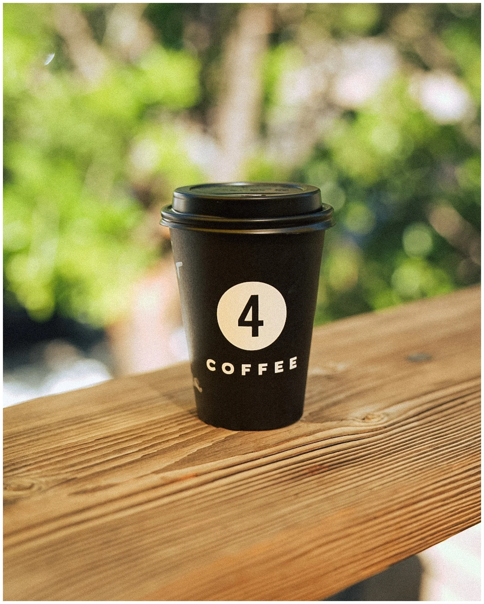 Black coffee cup with number 4 and "COFFEE" label on wooden surface, outdoor blurred background.