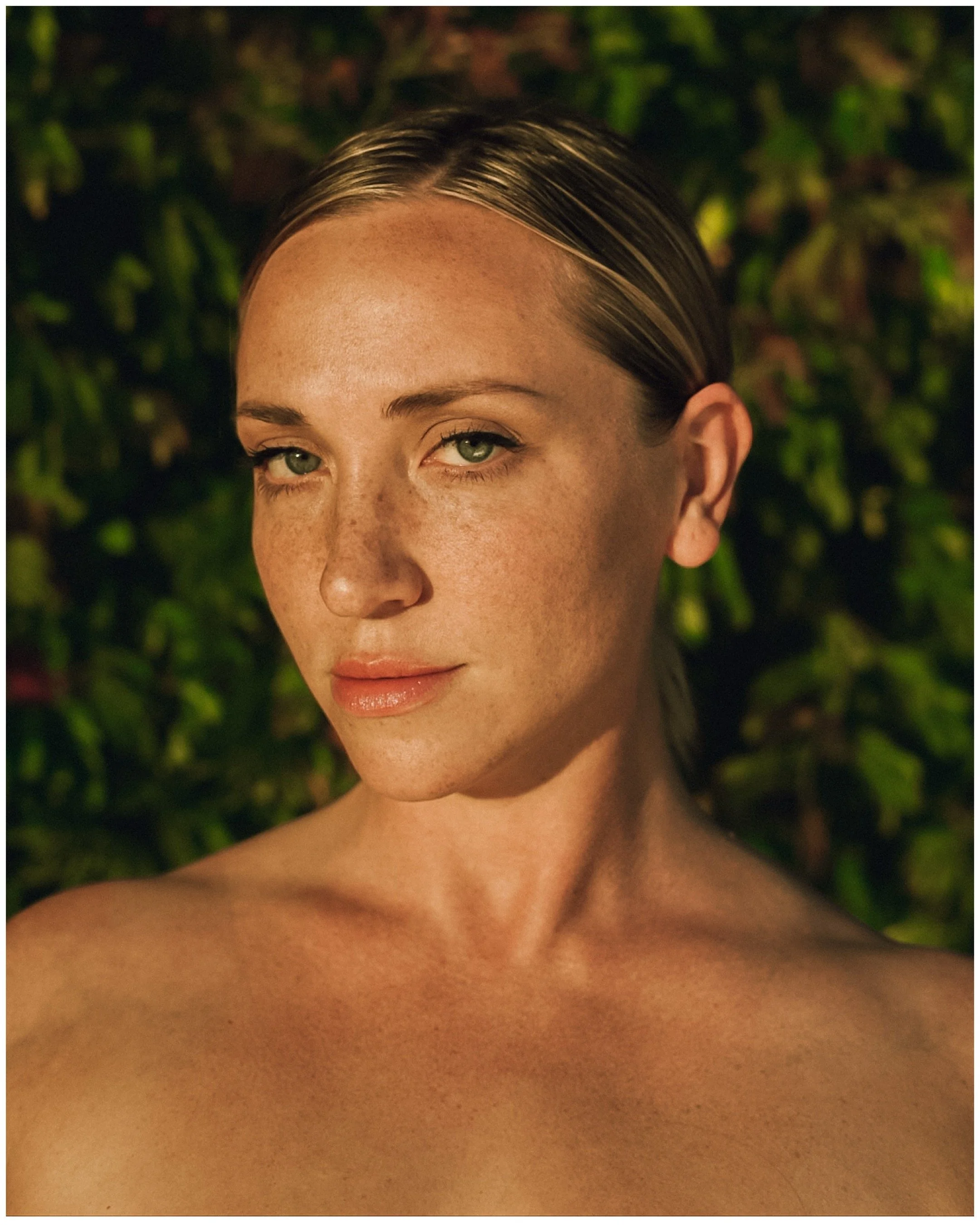 A person with light skin, freckles, and green eyes, standing in front of a leafy background.