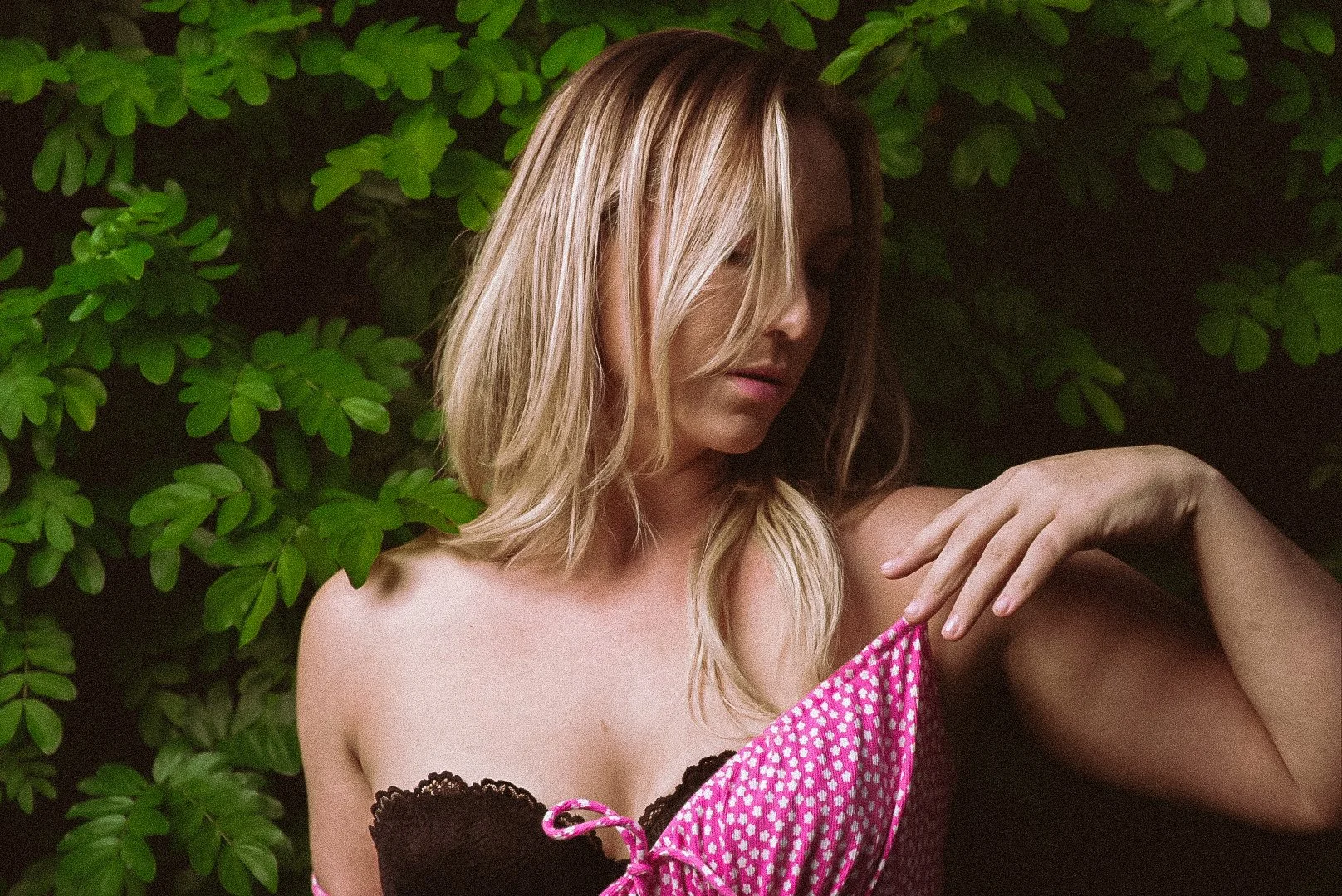 Woman with blonde hair wearing a pink and brown outfit, surrounded by green leaves.