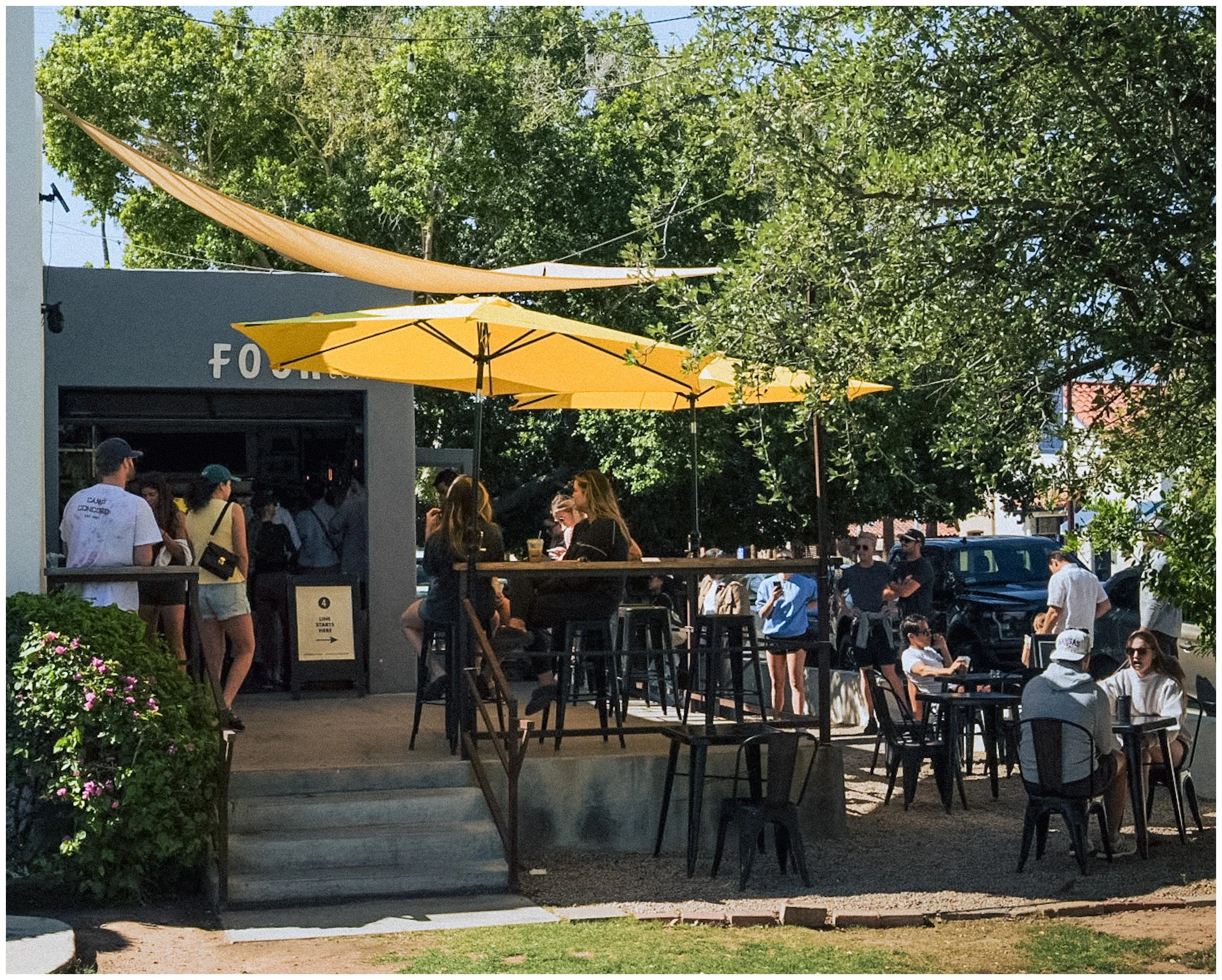 Outdoor cafe with yellow umbrellas and people seated at tables, surrounded by trees.