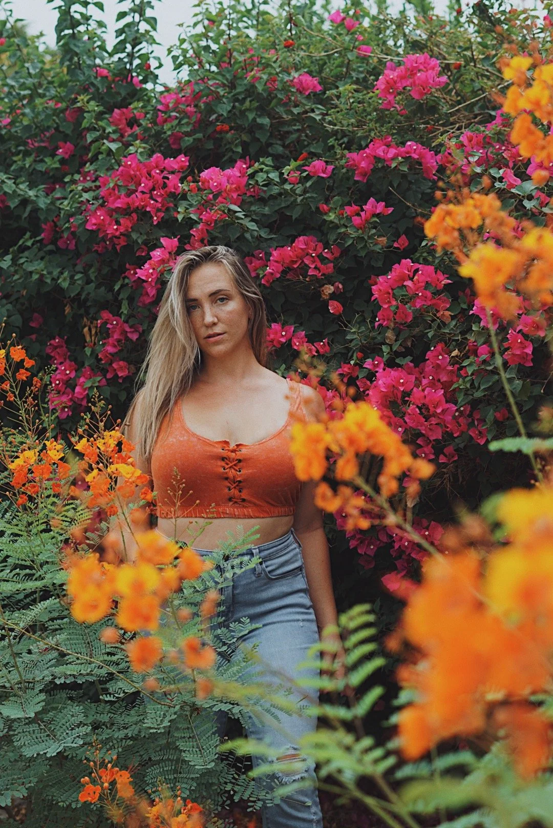 Person in an orange top and jeans standing amidst vibrant pink and orange flowers.