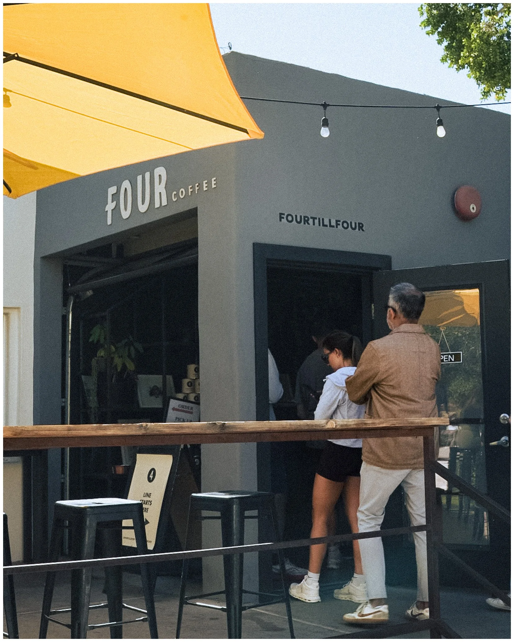 People entering a small coffee shop named Four Coffee, with outdoor seating and a yellow canopy.
