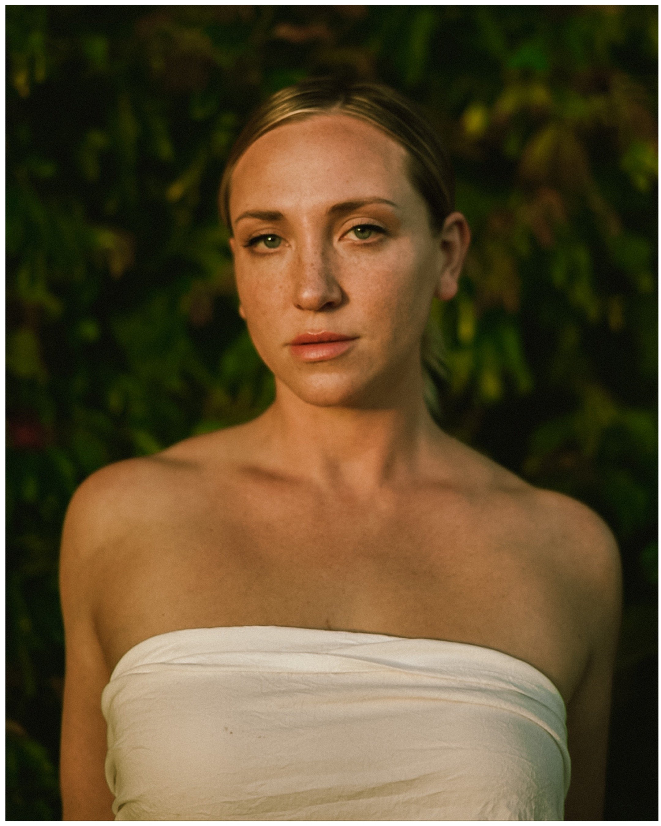 A woman with a neutral expression wearing a white strapless top, set against a dark, blurred foliage background.