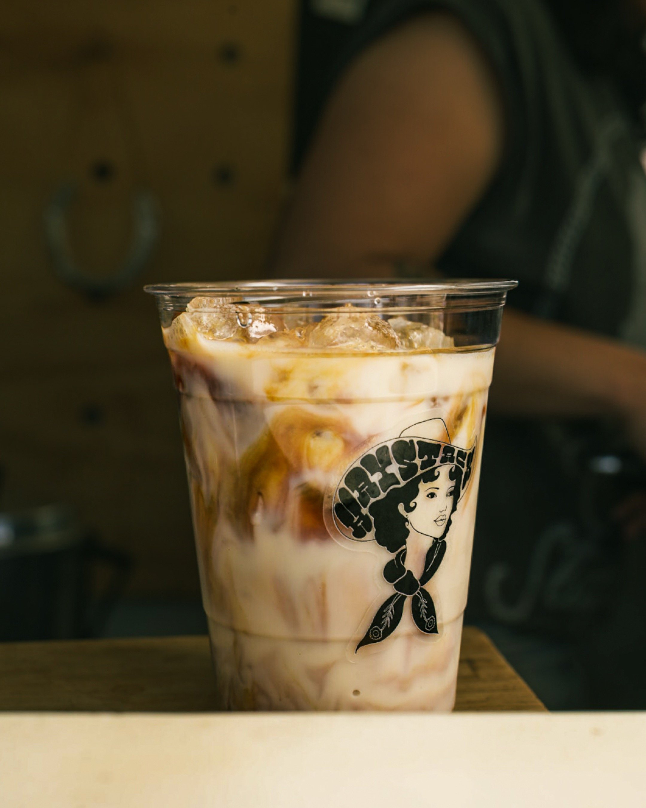Iced coffee in a clear cup with a vintage-style logo of a woman wearing a hat and bandana.