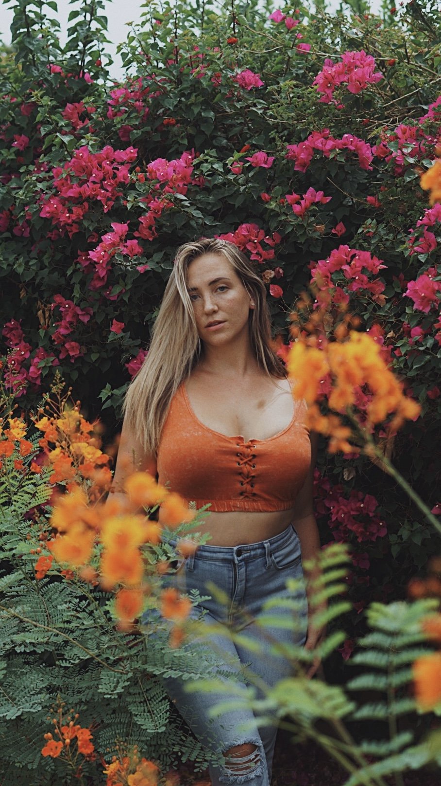 Woman in orange top and jeans standing among pink and orange flowers.