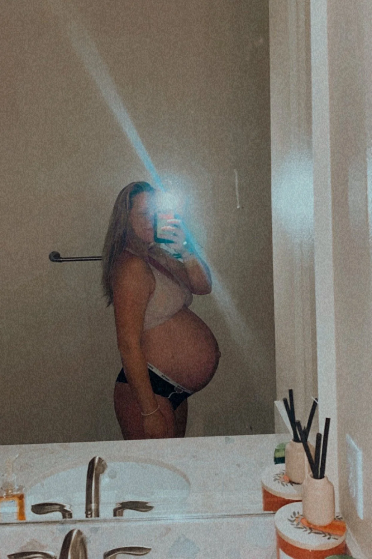 Pregnant woman taking a mirror selfie in a bathroom, wearing a beige bra and black underwear. Bathroom decor includes two small white vases with black reed diffusers, soap, and a mirror with a sink.