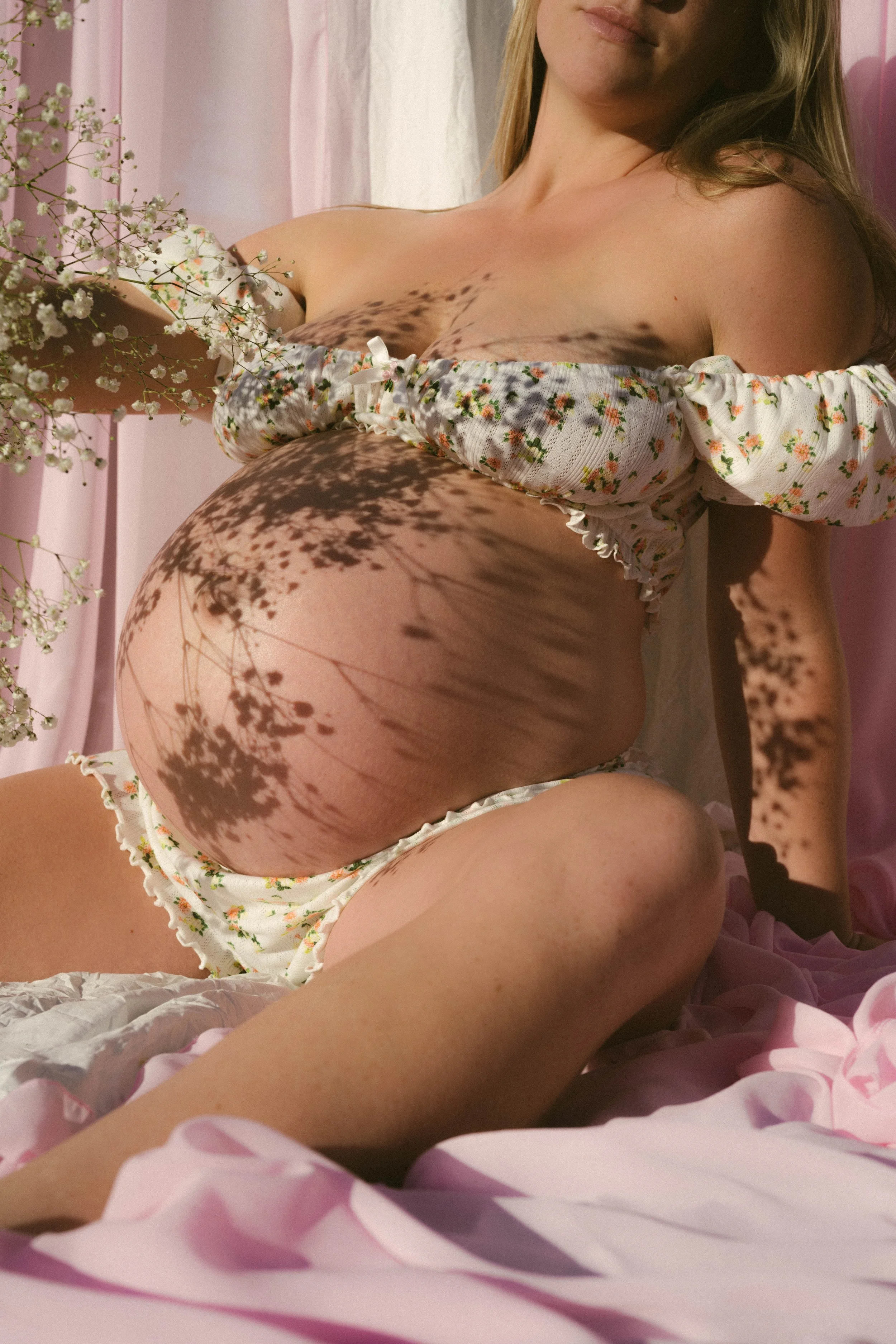 A pregnant woman in floral clothing sitting on a pink bed, with shadows of flowers cast on her belly and face.
