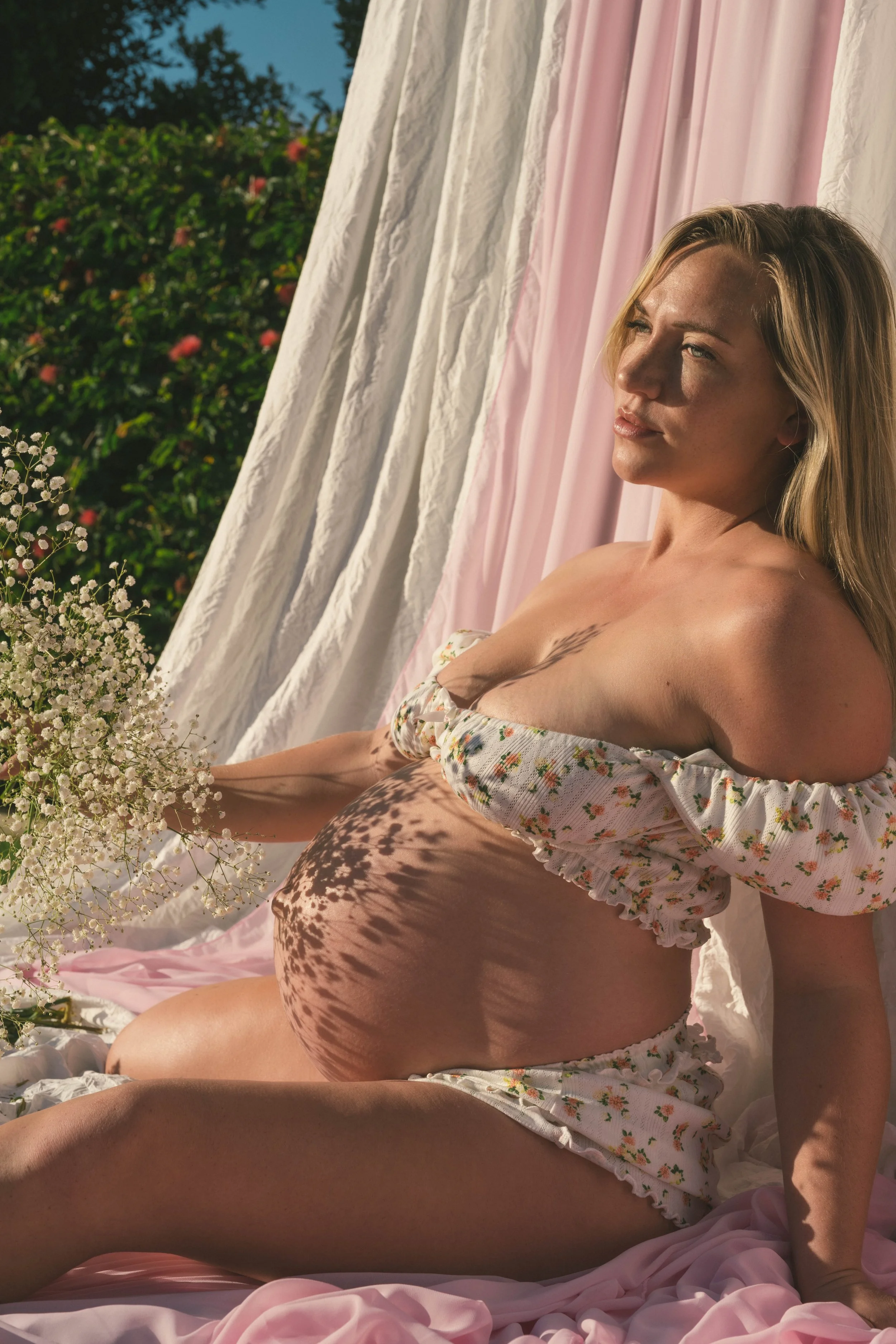 A pregnant woman with blonde hair sitting outdoors during daytime, surrounded by pink and white drapes and flowers, with sunlight casting shadows of leaves on her belly.