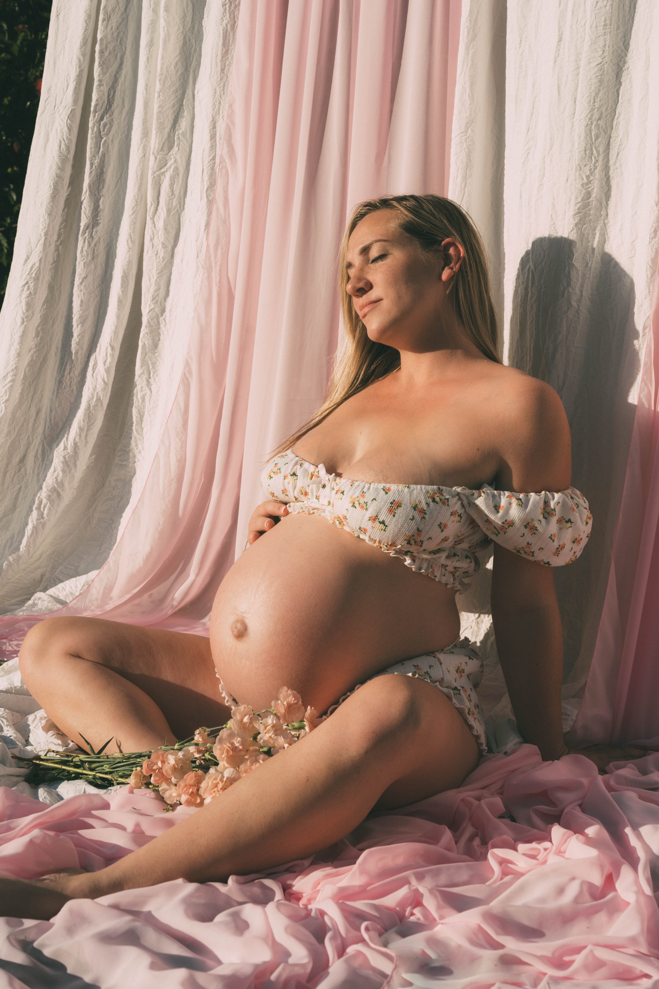 A pregnant woman with long blonde hair sitting on pink and white fabric, leaning against a background of pink and white curtains, with pink flowers placed on her lap and around her legs, smiling with closed eyes.