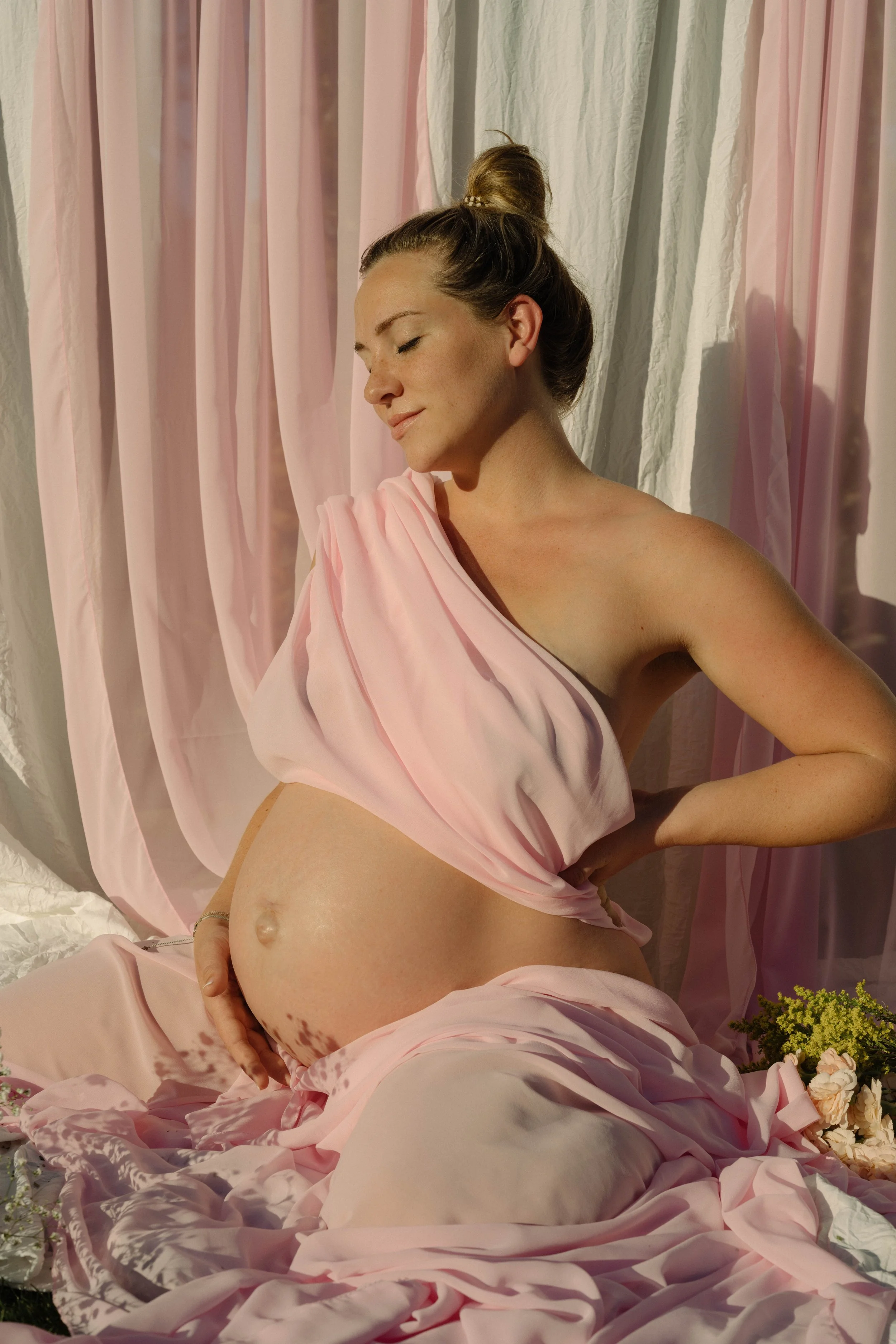 A pregnant woman with a bun hairstyle, wearing a pink satin fabric draped around her shoulders, sitting with her eyes closed, in a softly lit room with pink and white curtains and flowers around her.