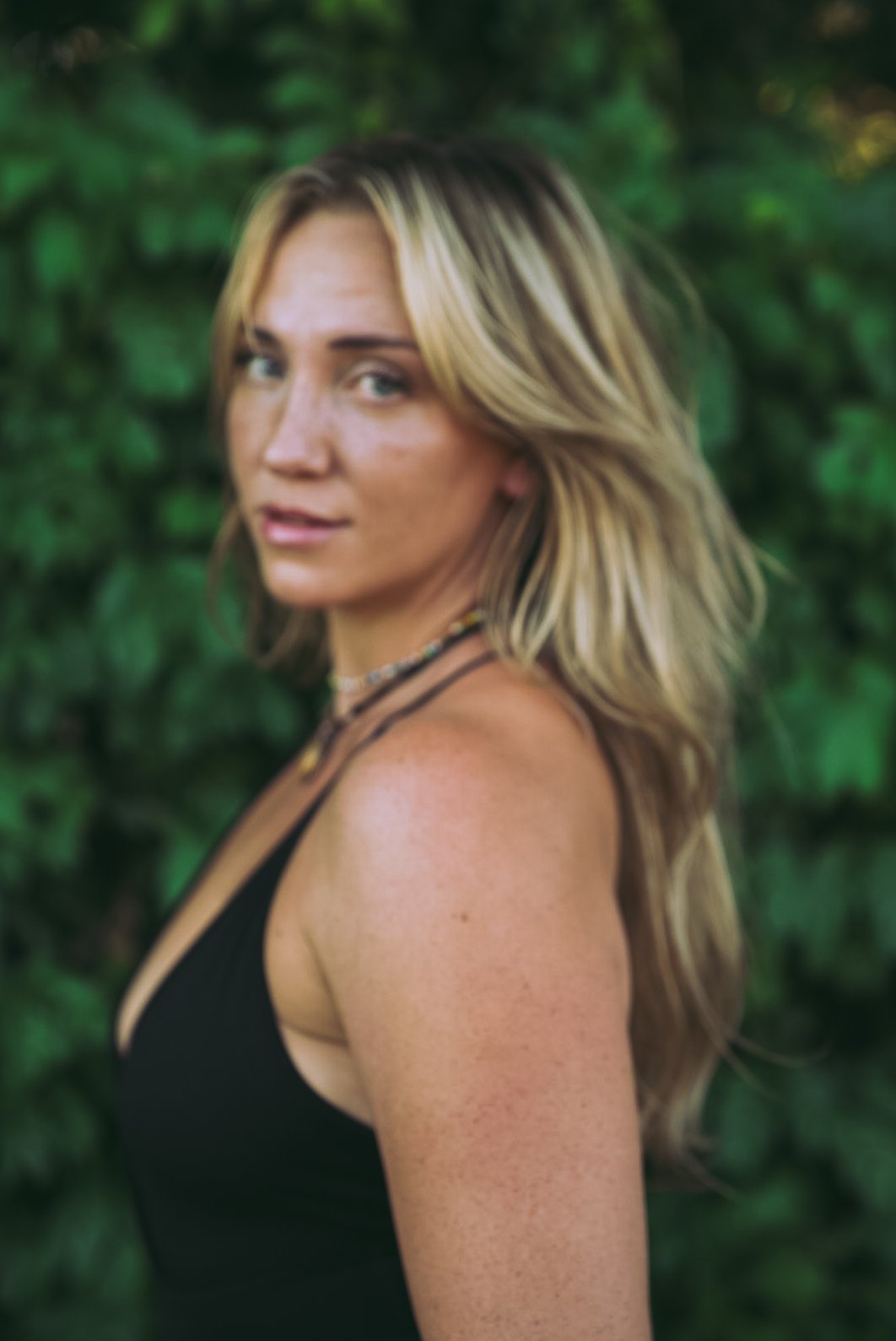 A woman with blonde hair and light skin wearing a black top, standing outdoors with green foliage in the background.