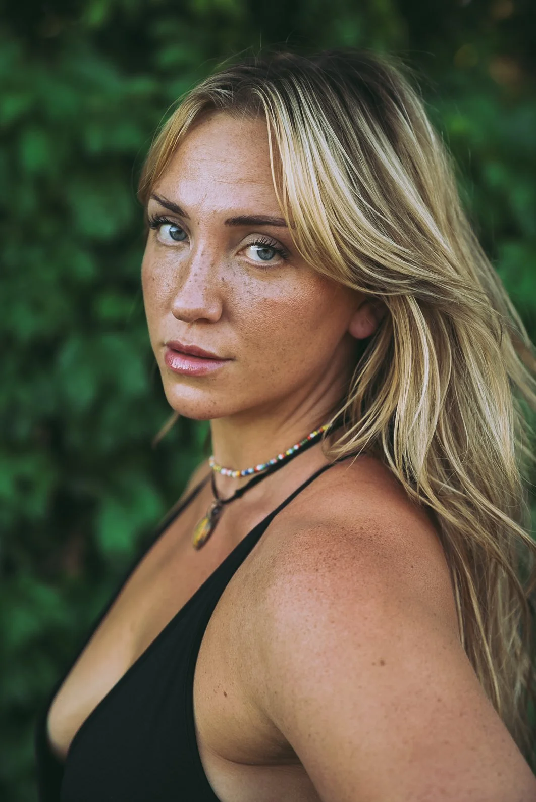 Close-up of a woman with blonde hair, blue eyes, freckles, and a black top, standing outdoors against a green leafy background.