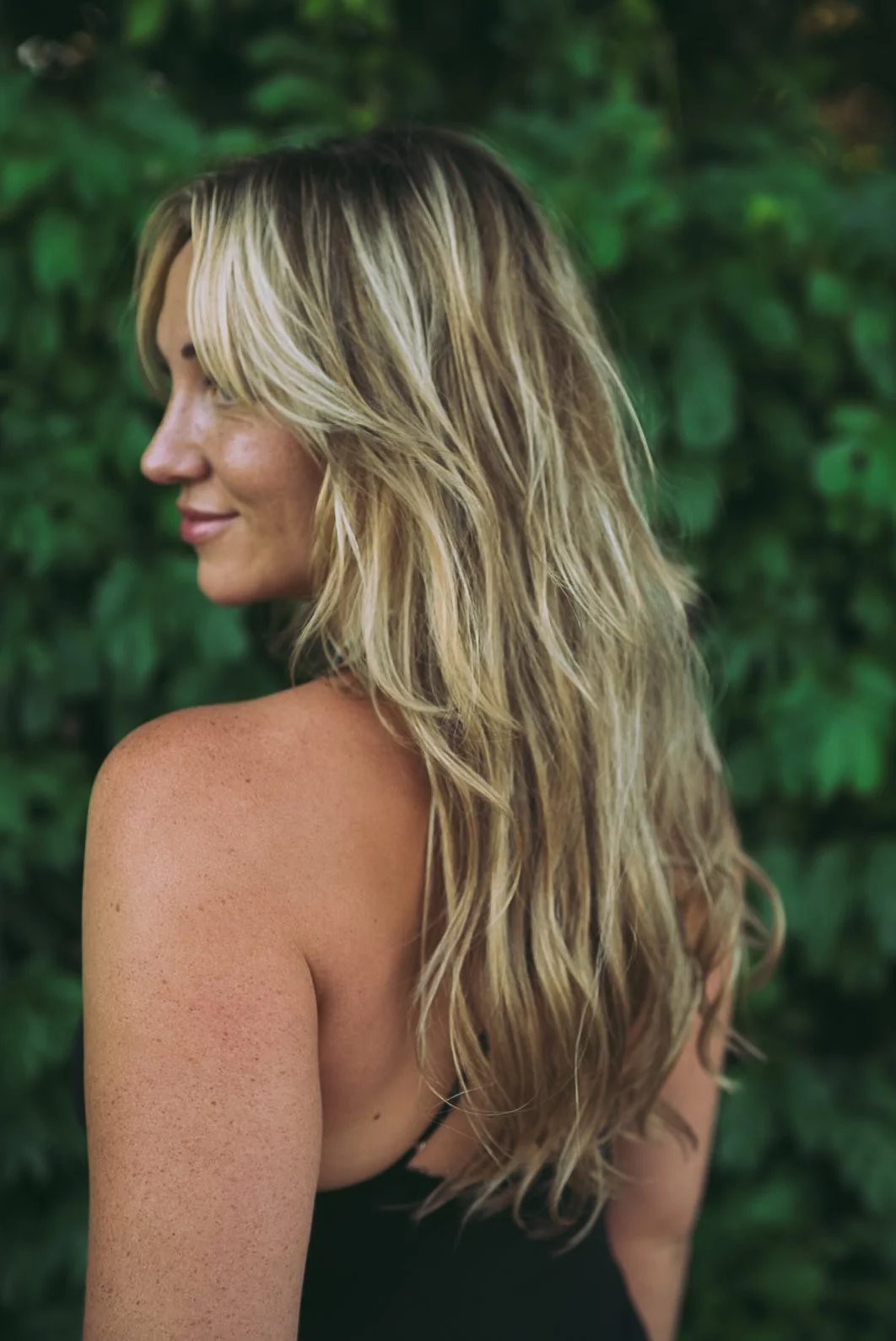 A woman with long, wavy blonde hair and tan skin standing outdoors with green foliage in the background, looking to the side with a slight smile.