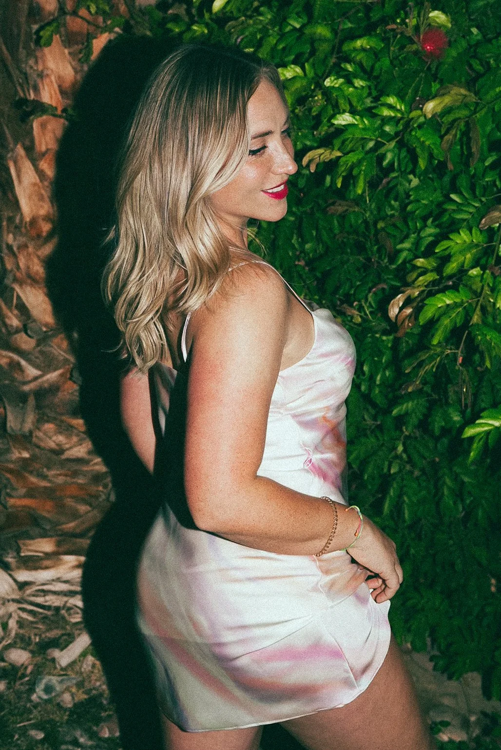 A woman with blonde, wavy hair smiling with her eyes closed, wearing a satin slip dress, standing outdoors against a backdrop of green foliage and a stone wall.