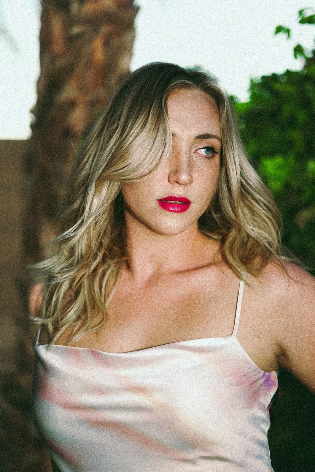 A woman with blonde hair, red lipstick, wearing a satin slip dress, looking to the side outdoors with trees in the background.