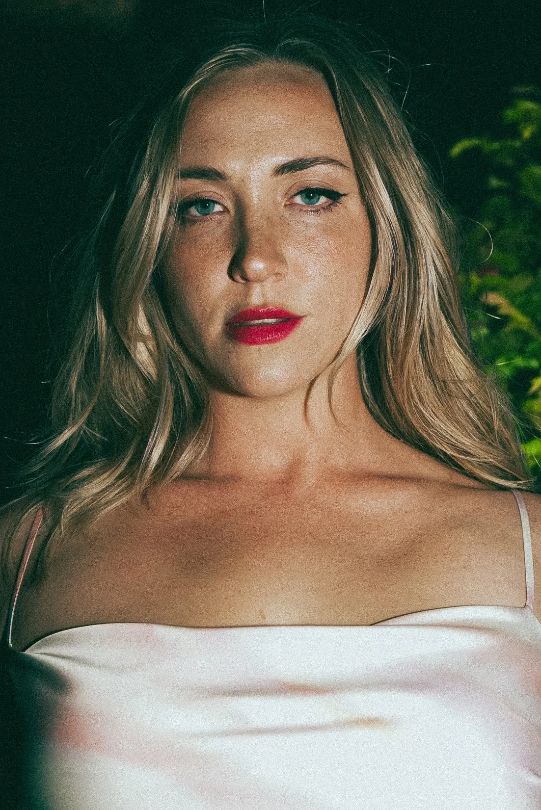 Close-up portrait of a young woman with blonde hair, blue eyes, red lipstick, wearing a white satin top, with a blurred greenery background.
