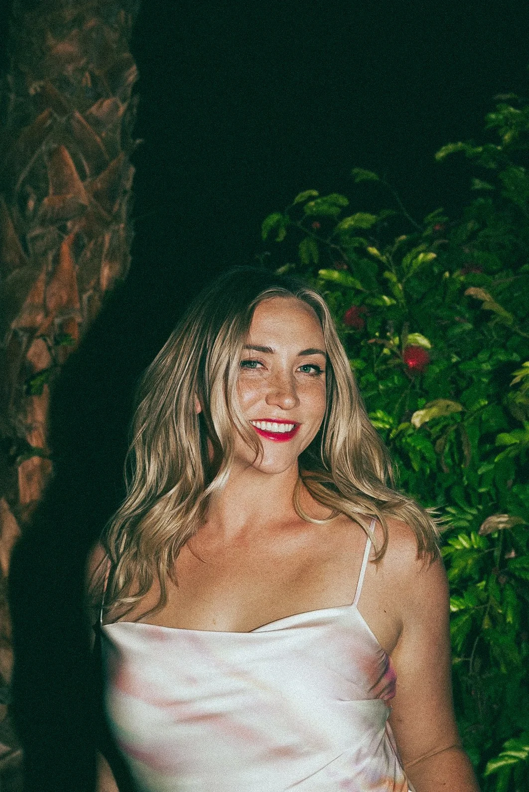 Smiling woman with long wavy blonde hair wearing a light pink satin dress, standing outdoors at night near greenery and a palm tree.