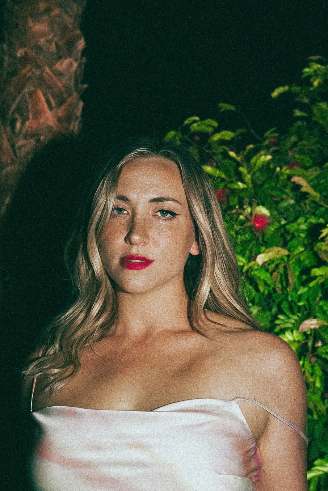 A woman with long blonde hair, wearing a white satin off-shoulder top, with a background of green plants and a stone wall.