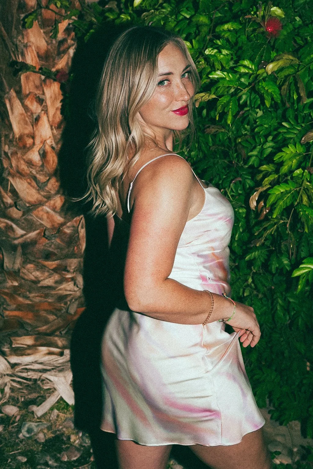 A woman with blonde wavy hair, red lipstick, wearing a silk slip dress with pastel colors, standing outdoors near a tree and green foliage.