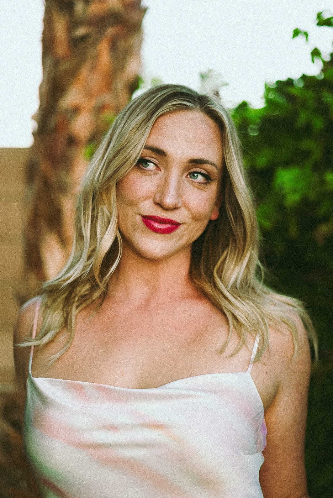 A woman with blonde hair and red lipstick, wearing a light pink spaghetti strap top, stands outdoors near a tree with greenery in the background.