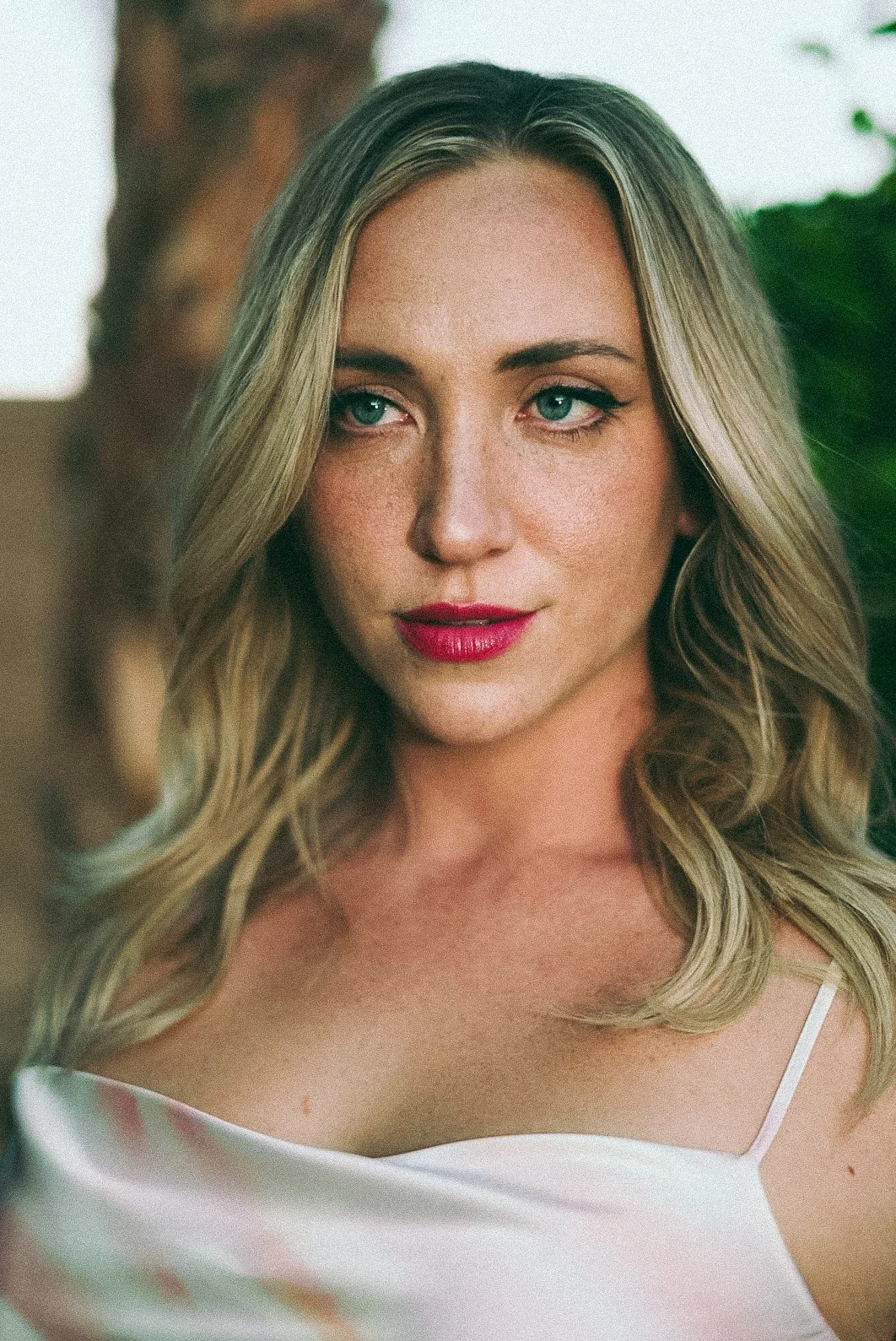 A woman with blonde hair, blue eyes, and red lipstick, wearing a white top with thin straps, standing outdoors with green foliage and a tree trunk in the background.