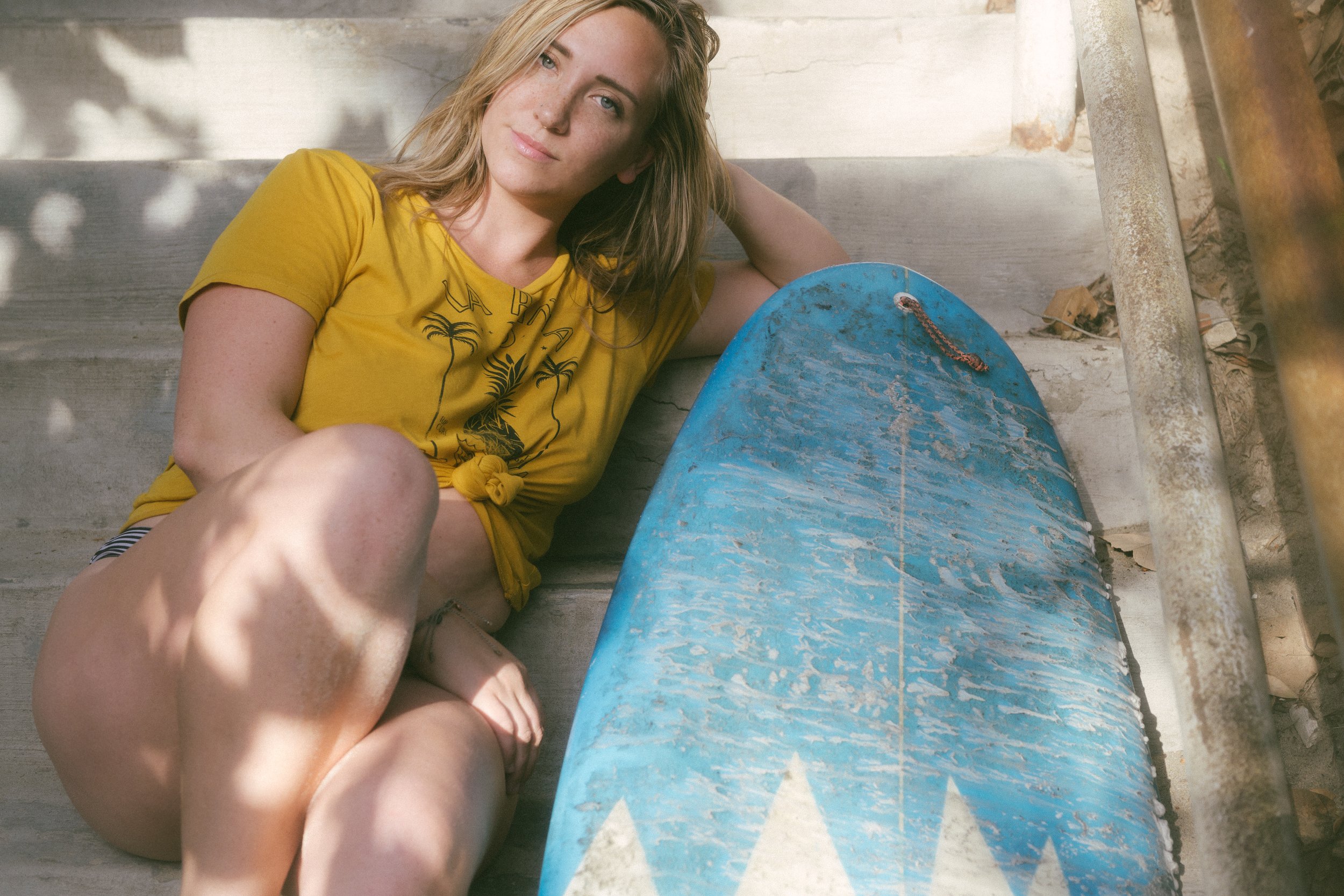 A young woman with long hair, wearing a yellow t-shirt, lying on a set of concrete stairs with a worn blue skateboard beside her.