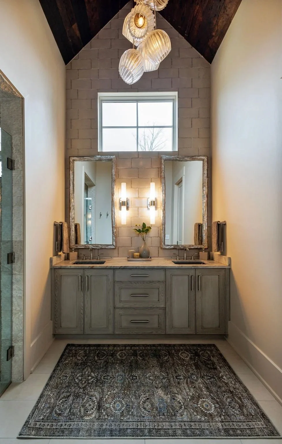 A modern bathroom vanity with two wood-like resin framed mirrors, a vase with green leaves and bath accessories, and two glowing wall sconces, with a window and an abstract blown glass chandelier hanging from a wooden vaulted ceiling.