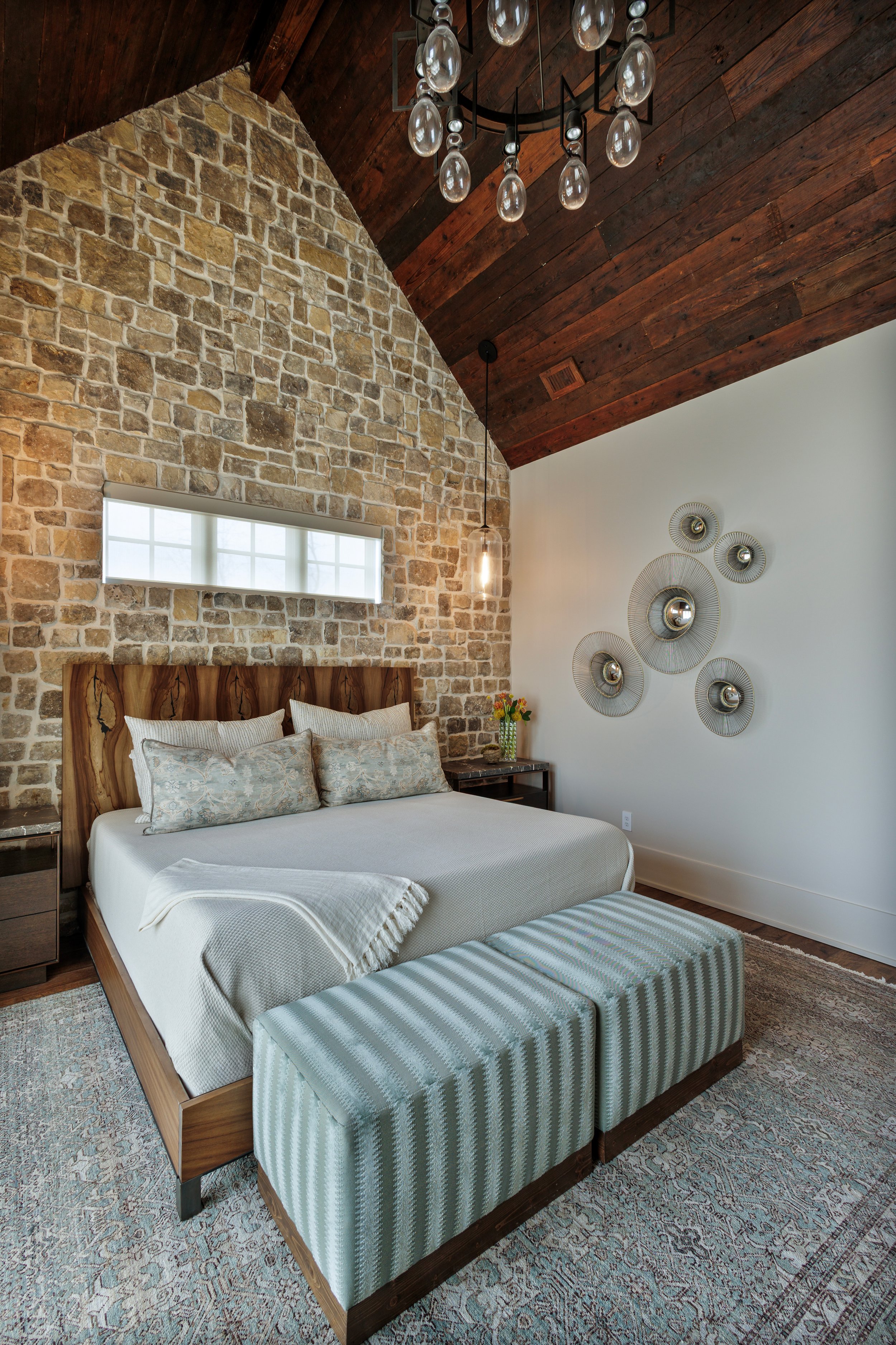A rustic primary bedroom with a stone wall behind the bed, a textural wood vaulted ceiling, and metal mirror wall art & blue stools. The bed has a wooden live edge headboard & linen and blue colored bedding. A glass chandelier hangs from the ceiling.