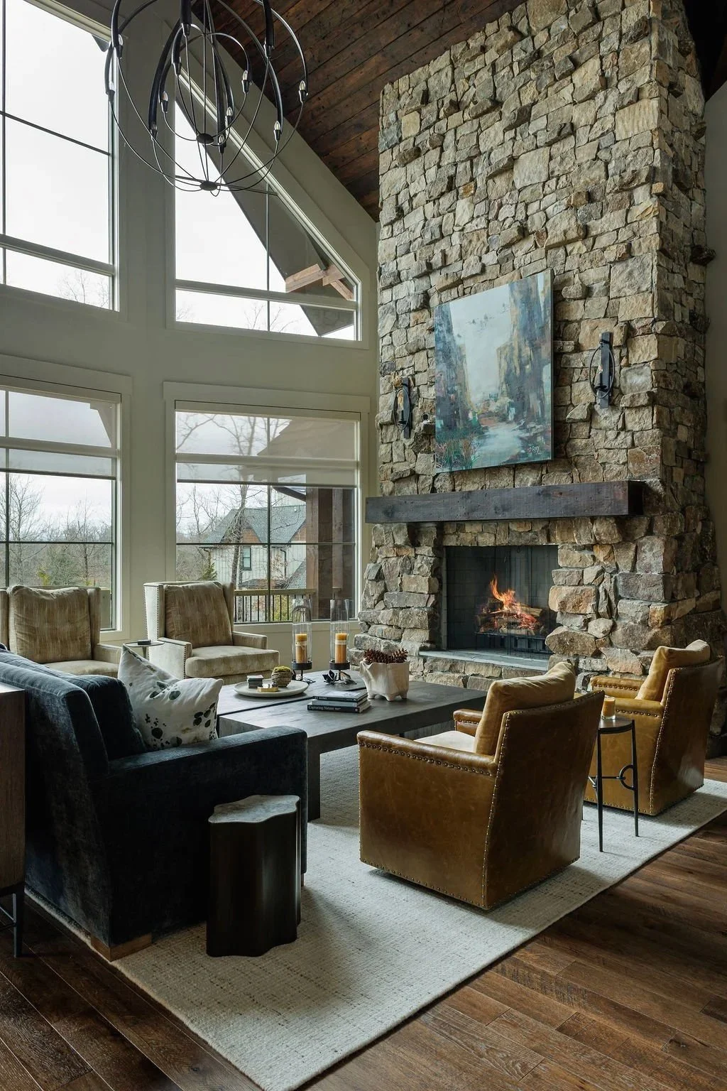 Living room with large windows and towering stone fireplace. The cozy living space features with charcoal sofa, ochre leather chairs & two coffee tables styled with decor and tall hurricane vases with candles. Large abstract art hangs over the mantle