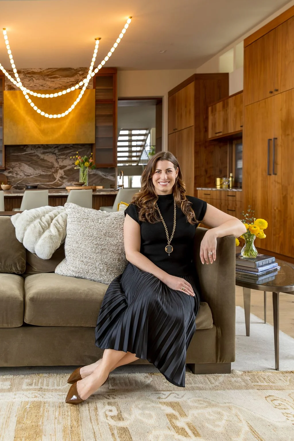 Principal Designer, Erin di Scipio, sits on an olive green sofa in a modern living room, smiling at the camera. Behind her is a contemporary kitchen with walnut cabinetry, brass hood and glowing swagged pendants made of strands of alabaster.